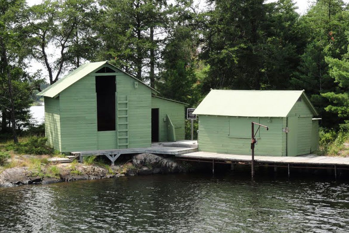 Two light-green wooden structures sit on a wooden dock over a calm lake, surrounded by dense green trees.