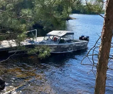 A camouflage-patterned motorboat is tied to a wooden dock on a calm, sunny lake, surrounded by pine trees.