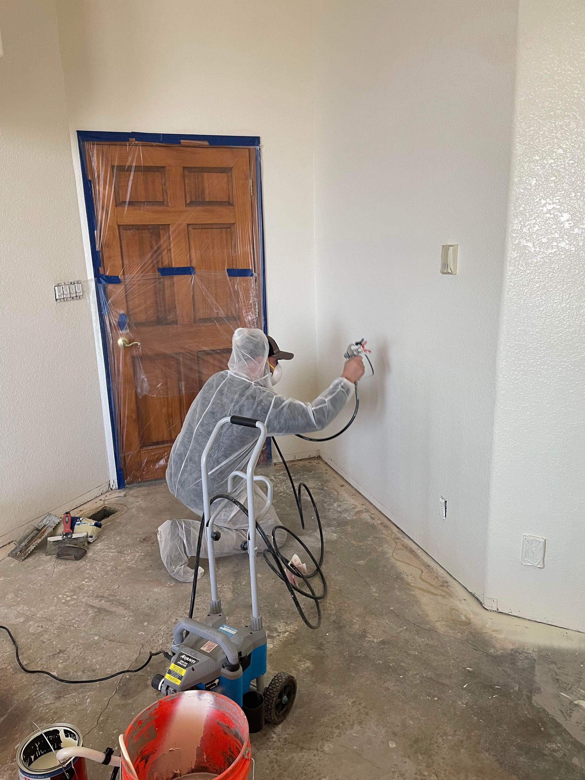 Person in protective suit spraying a white textured finish on an interior wall with a paint sprayer.
