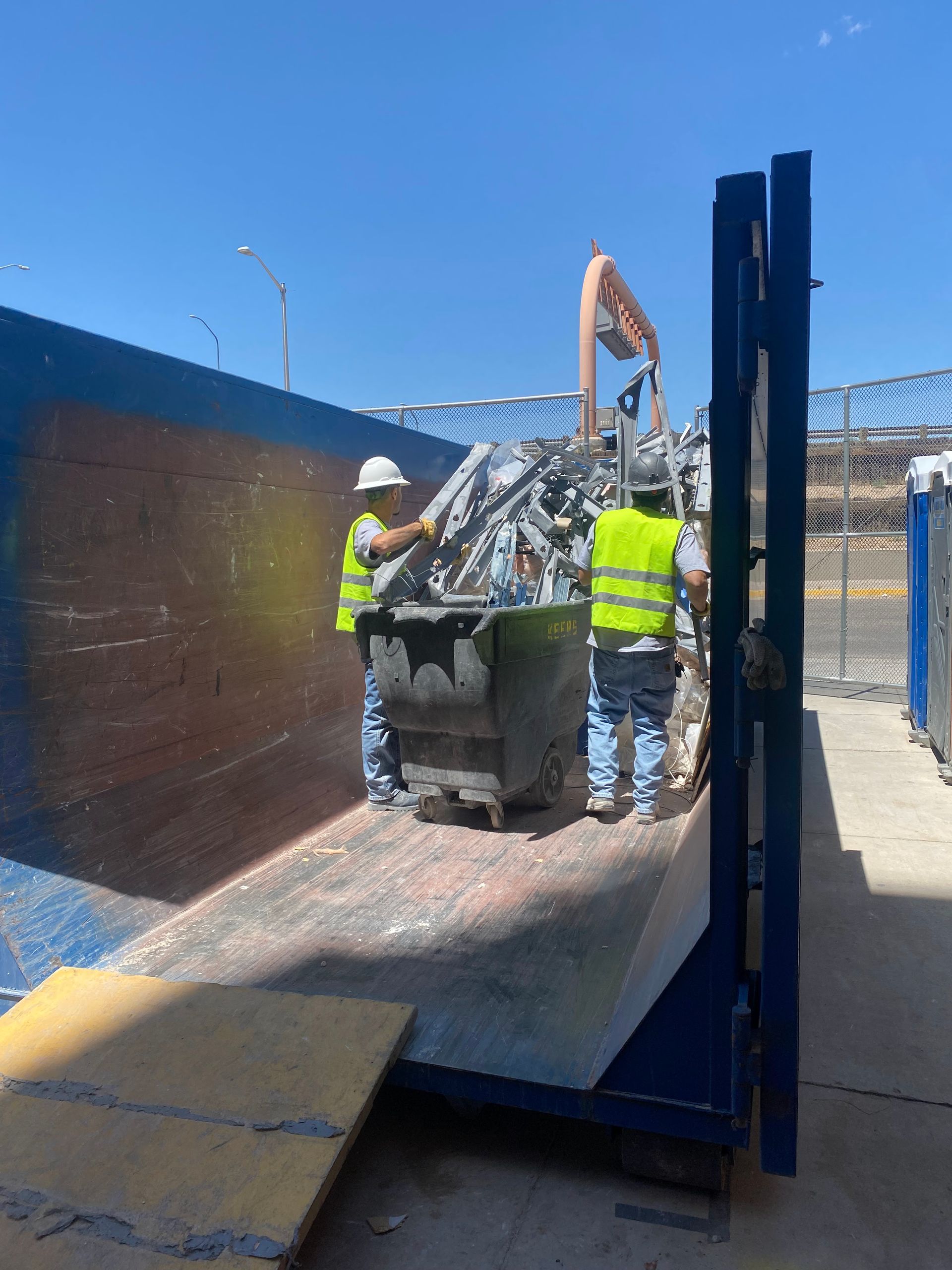 Two workers in safety vests and a mechanical arm loading scrap metal into a dumpster outdoors.