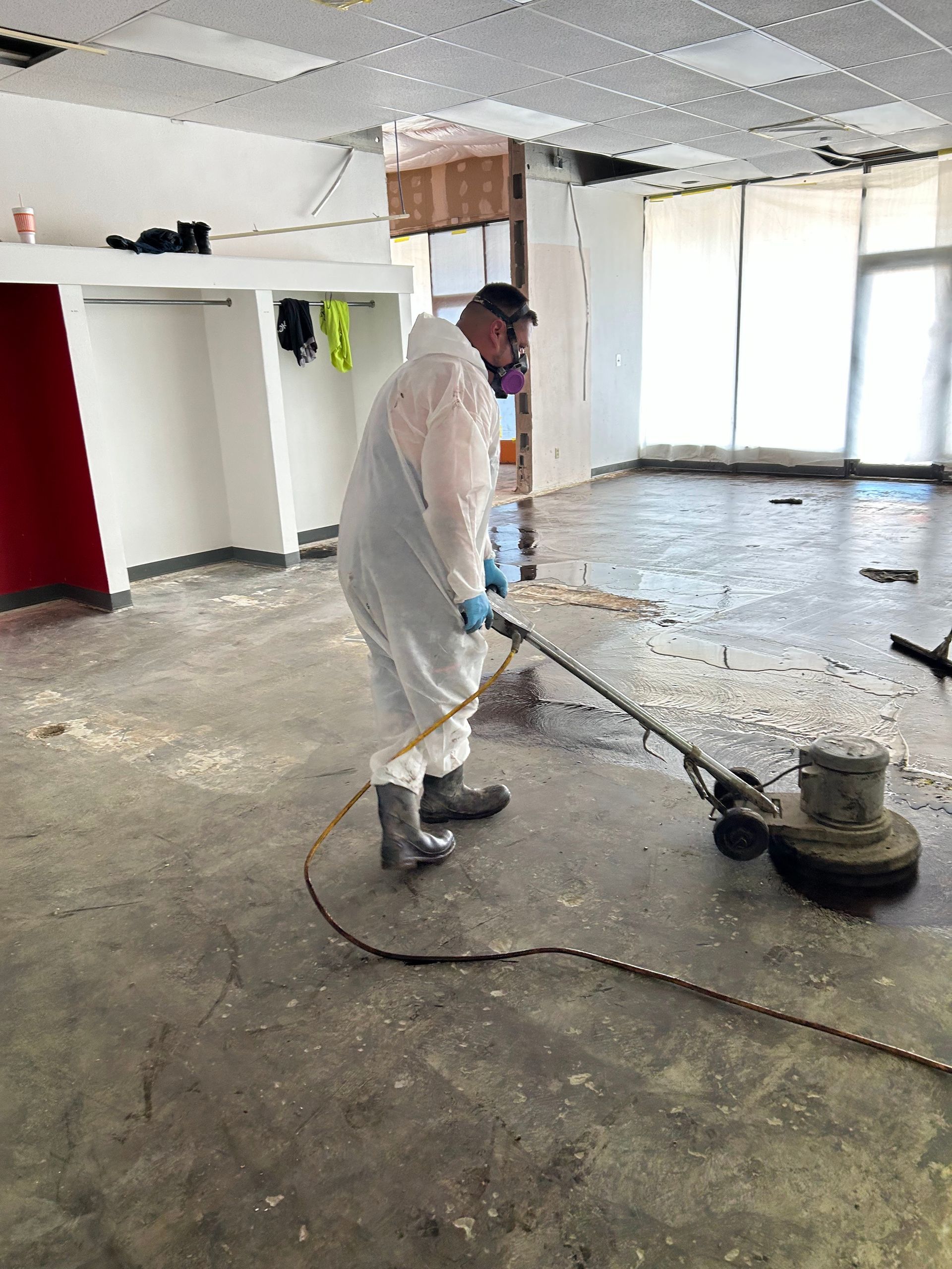 A person in protective suit, cleaning a concrete floor with a machine in an empty room.