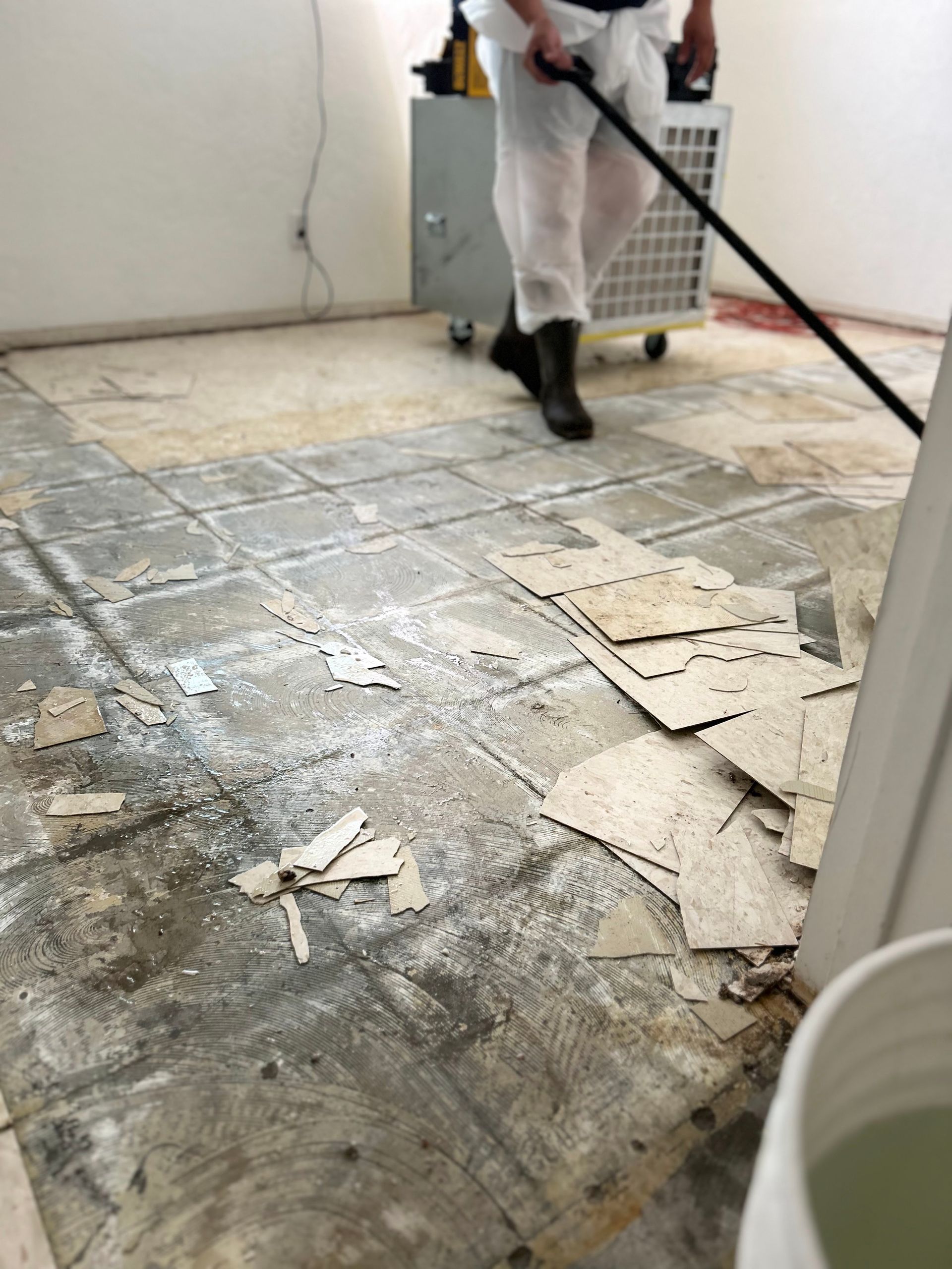 Person removing floor tiles, wearing protective gear. Debris and equipment on the floor.