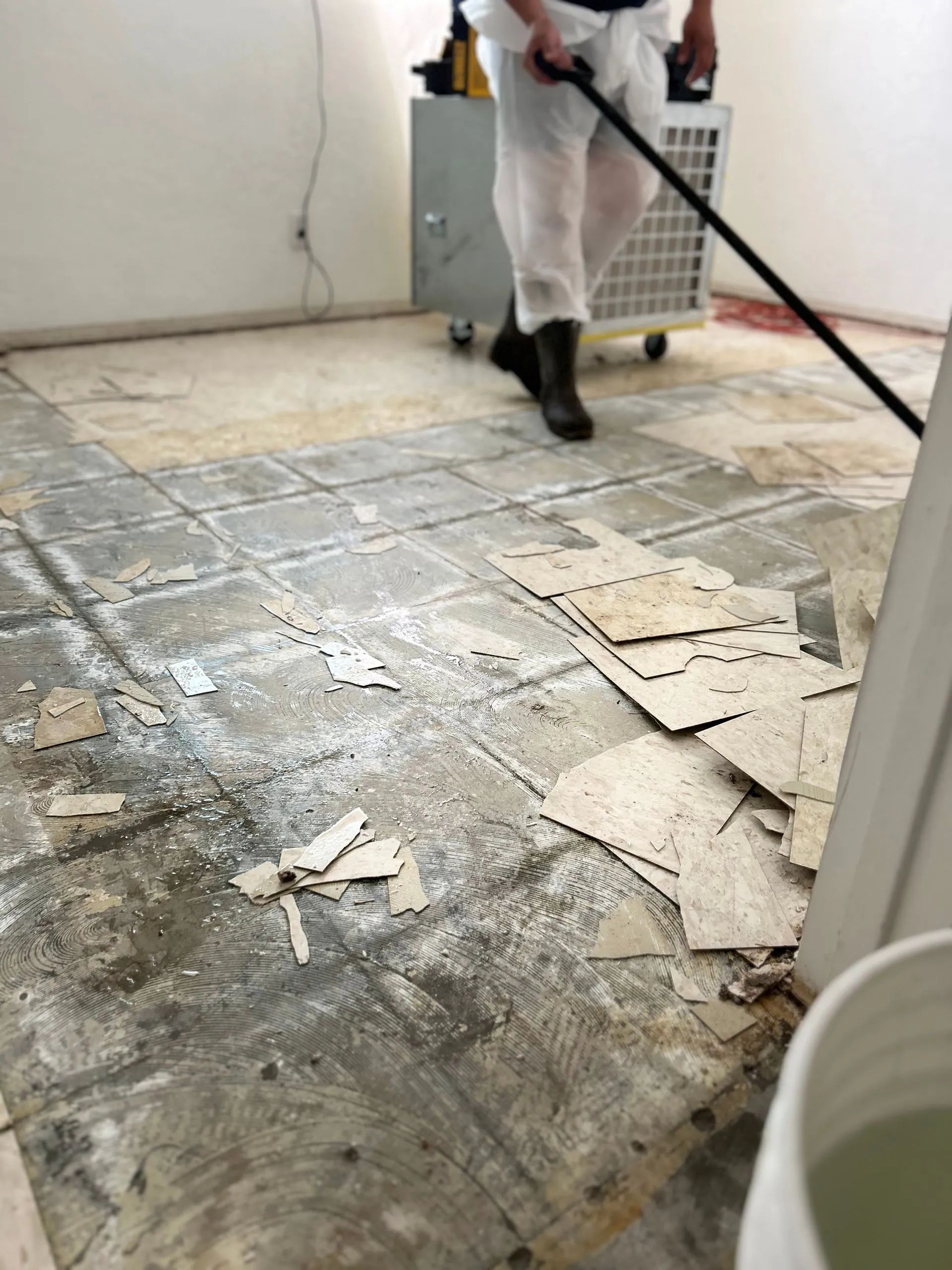 Person in white coveralls mopping a damaged floor with peeling paint in a sparse room.
