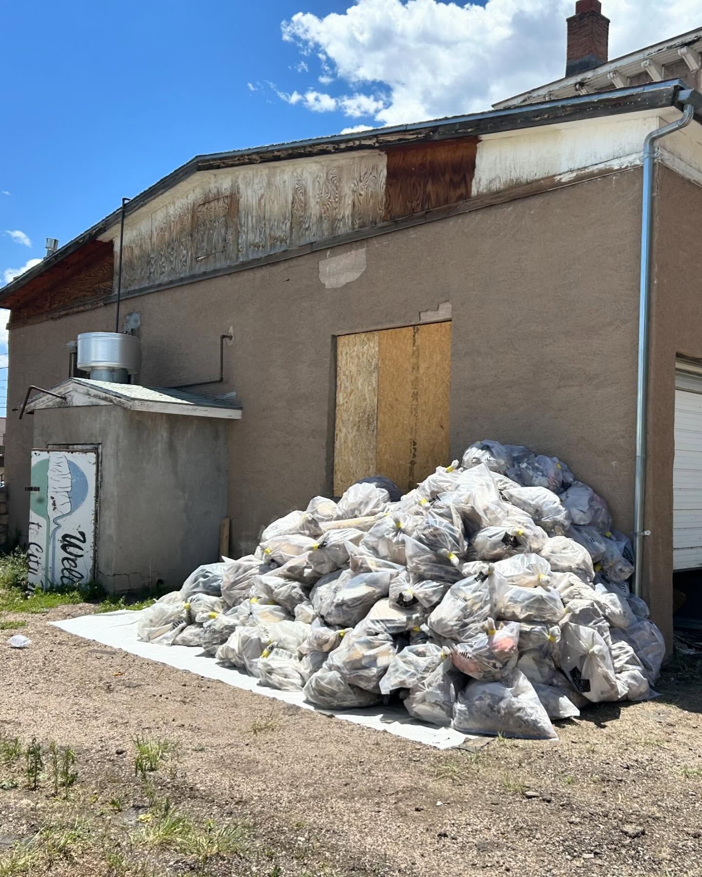 Exterior view of a tan building with a pile of bags. A small shed is to the left, with a garage door to the right.