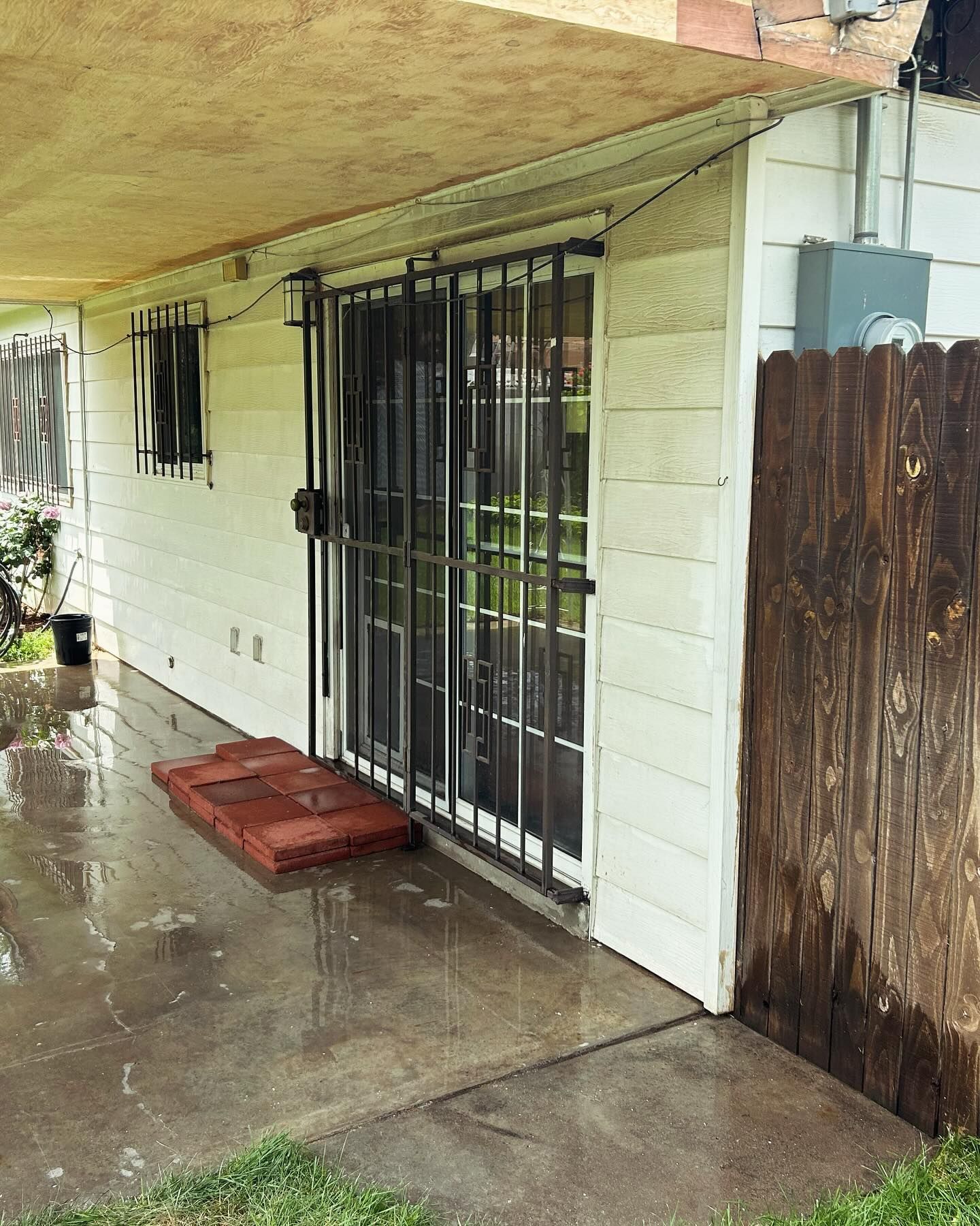 Sliding glass door with security bars, a concrete patio, and a wooden fence next to a white house.