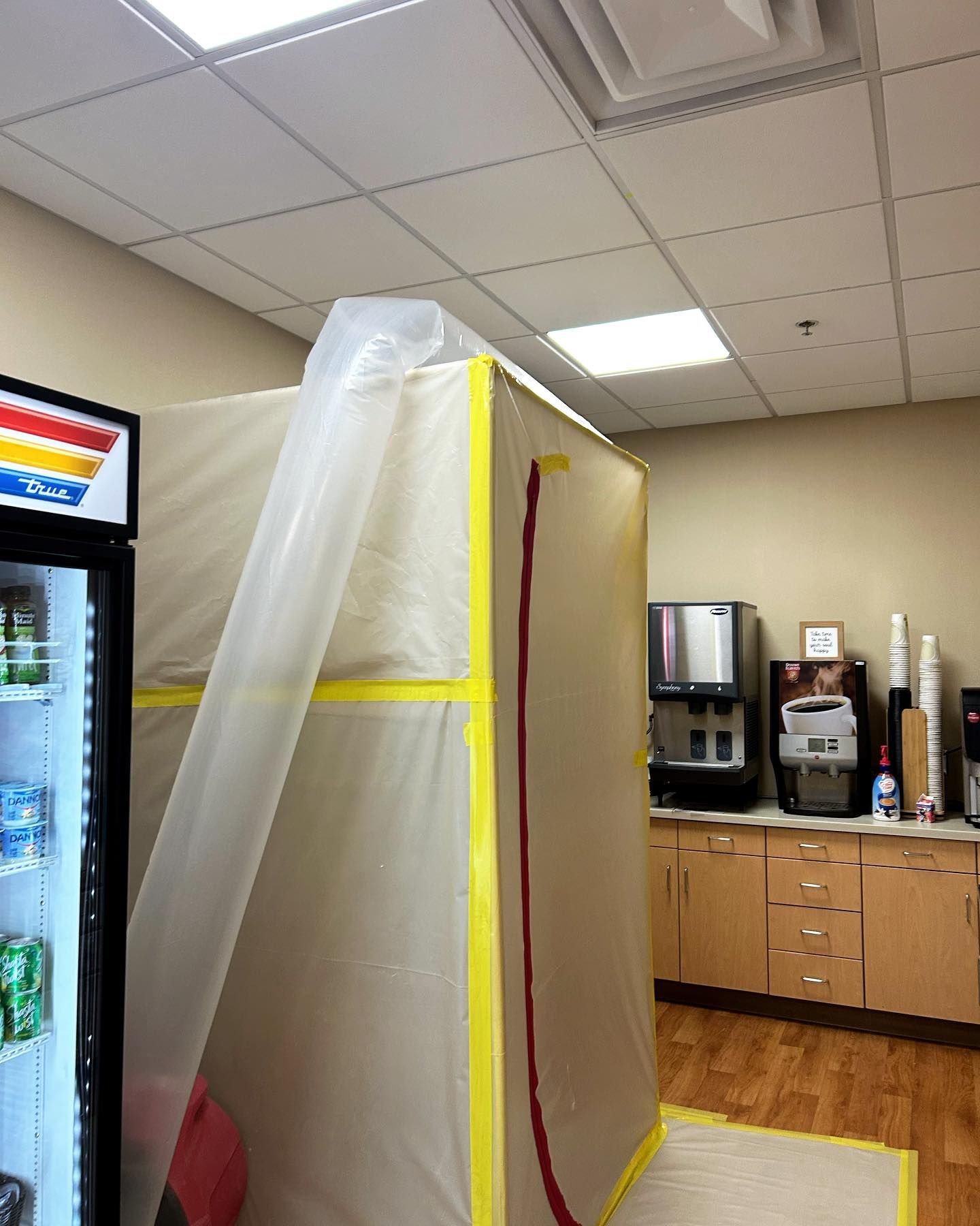 A refrigerator is covered in plastic sheeting and tape for repairs in a break room.