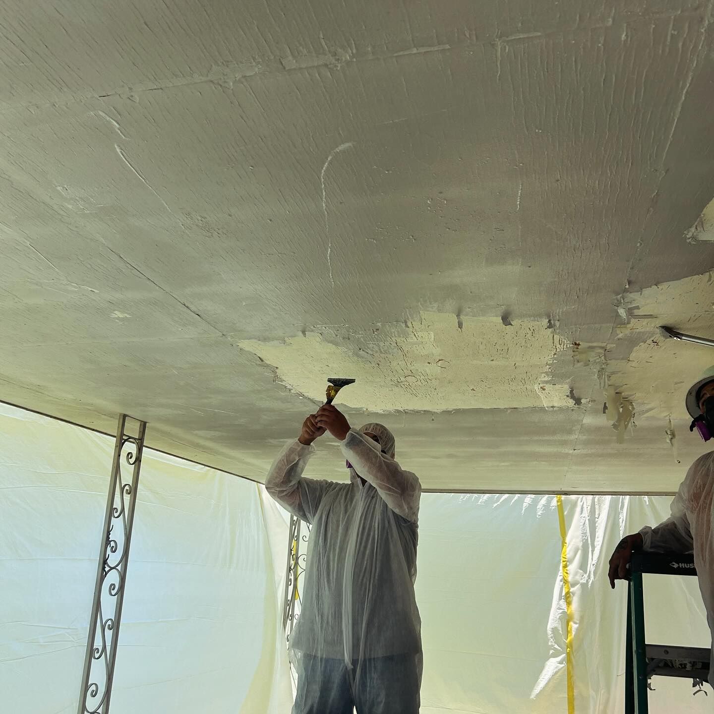 Person in protective suit scraping ceiling. White sheet barriers, sunny.