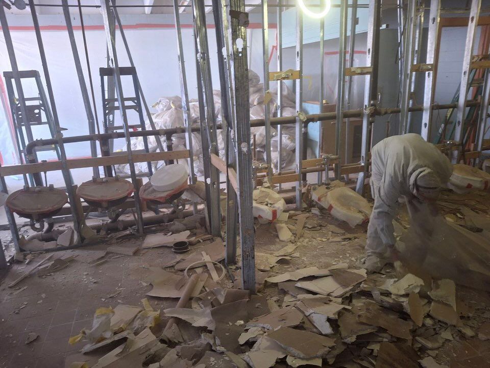 Interior construction site with exposed studs, debris on the floor, and a person working.