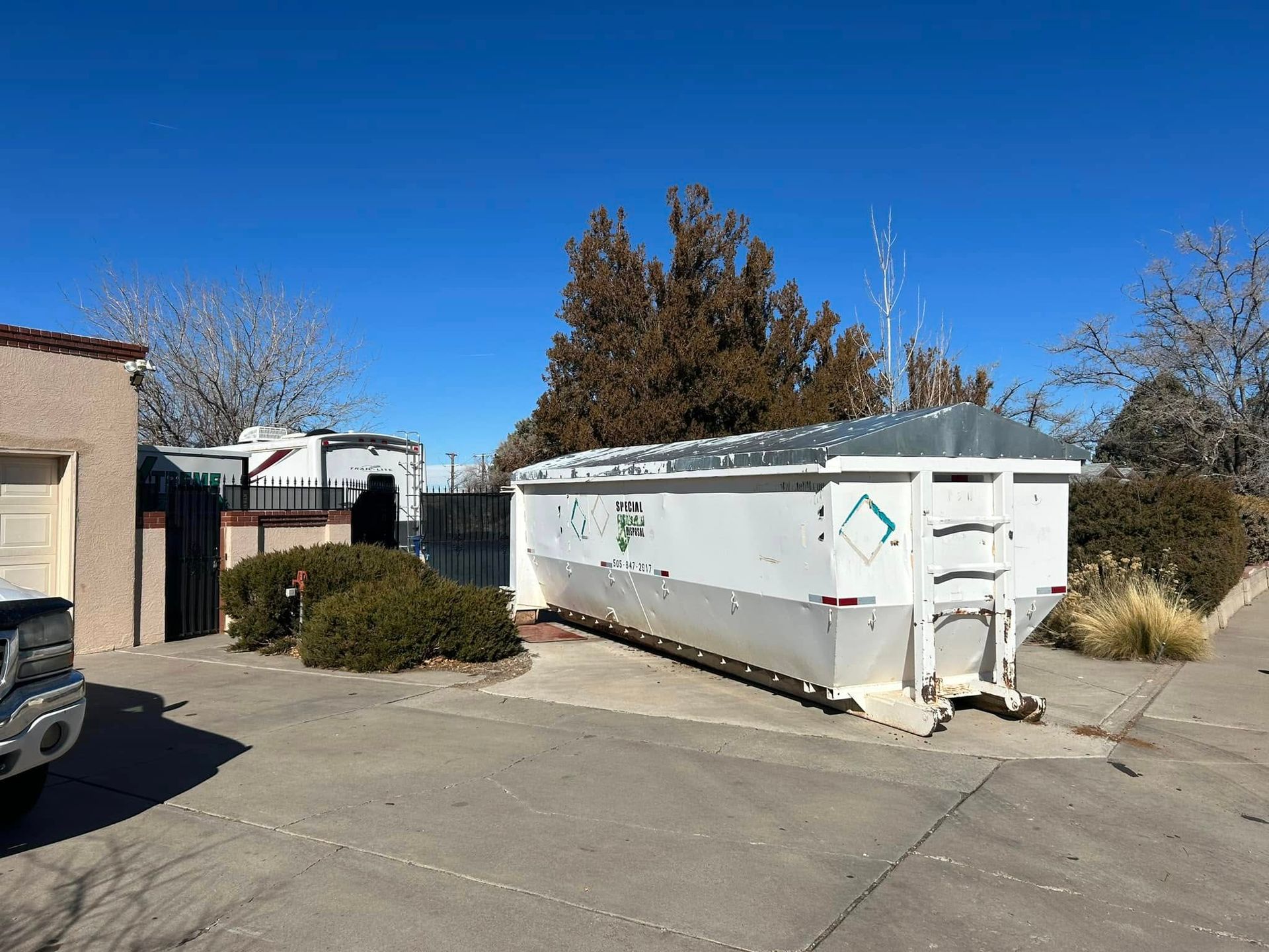 White dumpster on a paved surface, shrubs, trees, and buildings under a blue sky.