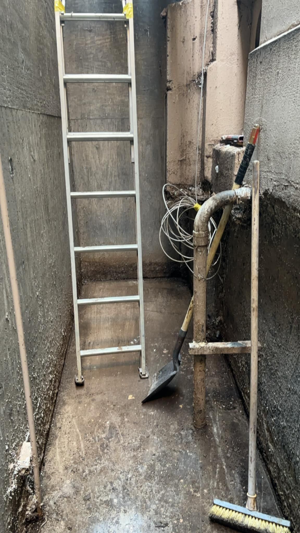 A ladder stands in a narrow, dirty alleyway with tools leaning against a pipe.