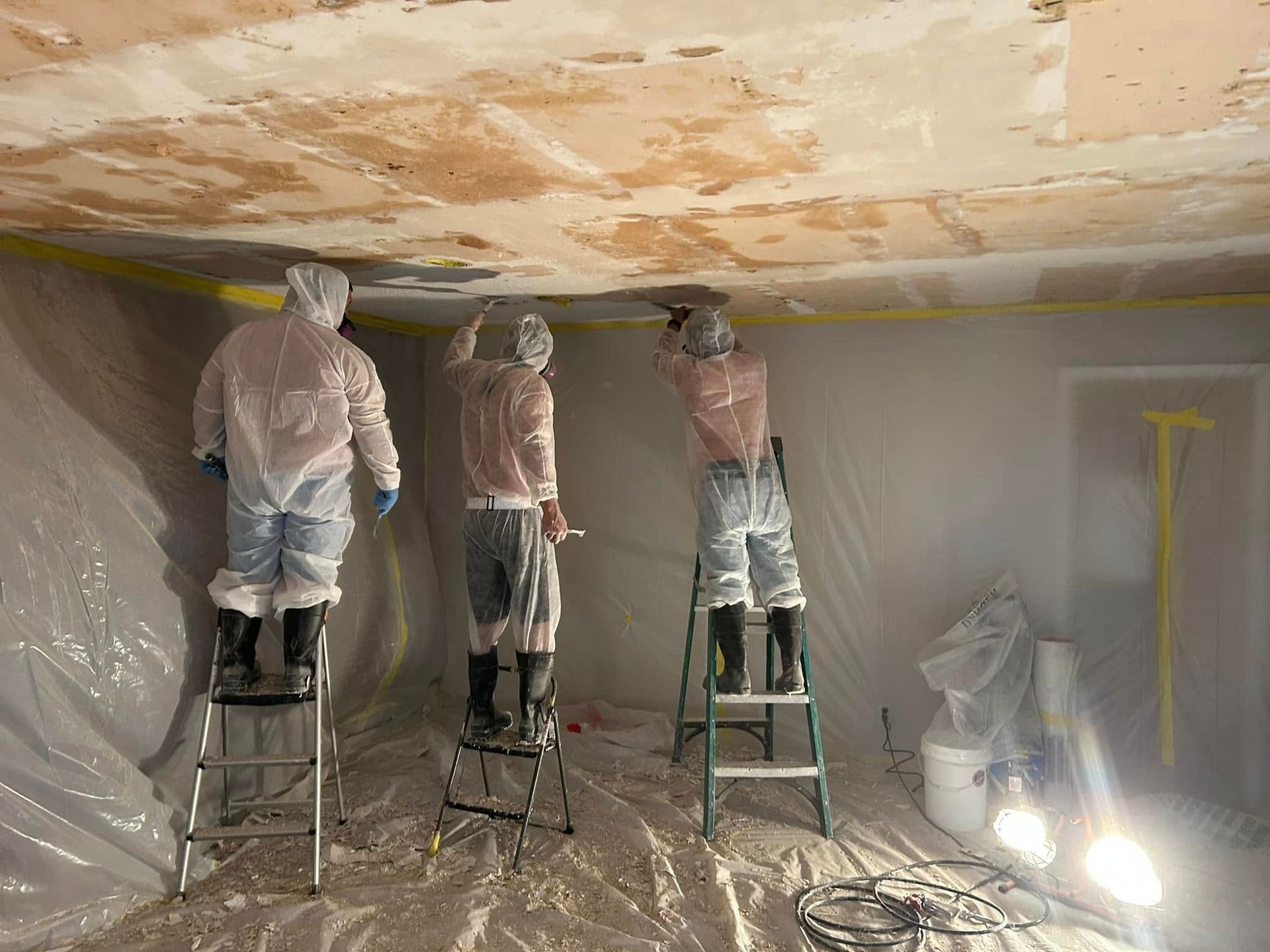 Three workers in protective suits remove ceiling material from a room.