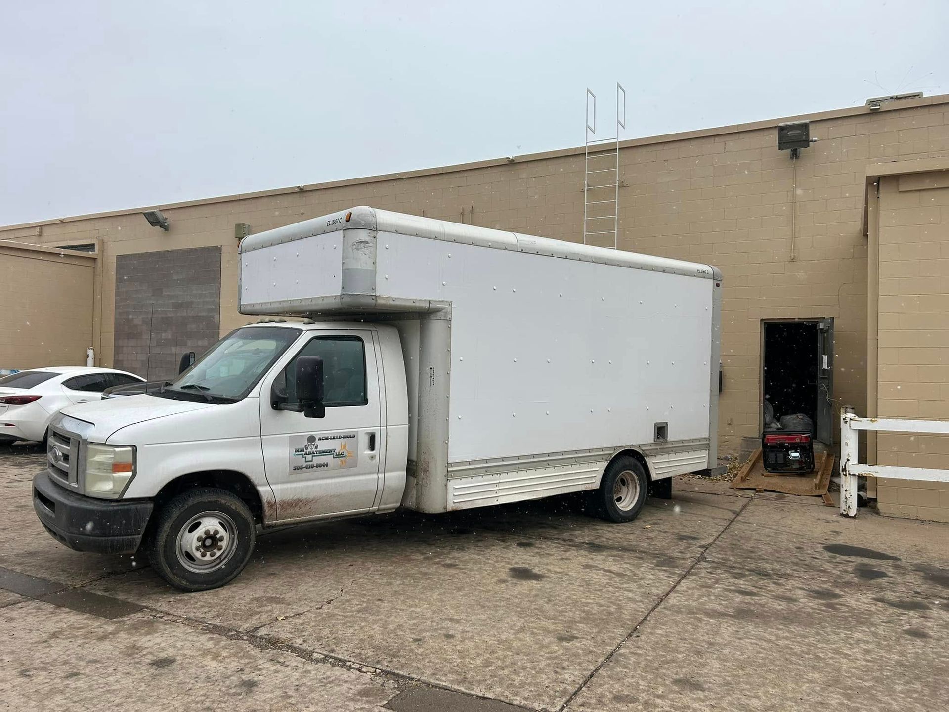 White box truck parked near a building with an open doorway. Snow falling.