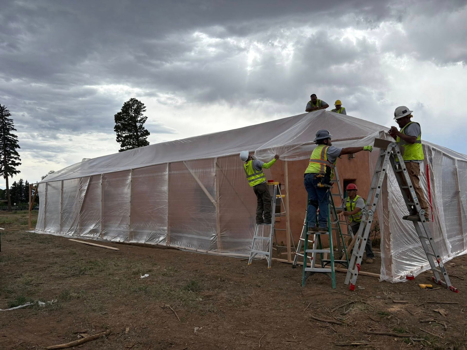 Construction workers on ladders installing clear plastic sheeting on a low, rectangular building, under a cloudy sky.