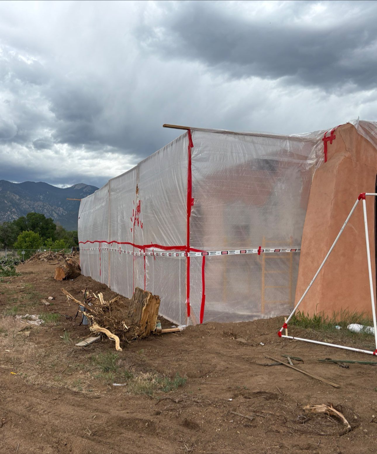 Clear plastic-covered structure secured with red tape, next to a reddish adobe building, under a cloudy sky.