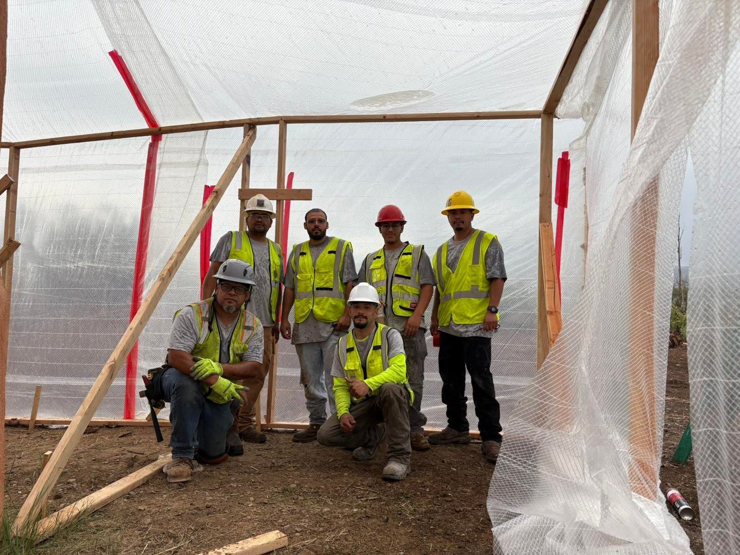 A group of construction workers pose in a makeshift shelter. They wear hard hats, vests, and work clothes.