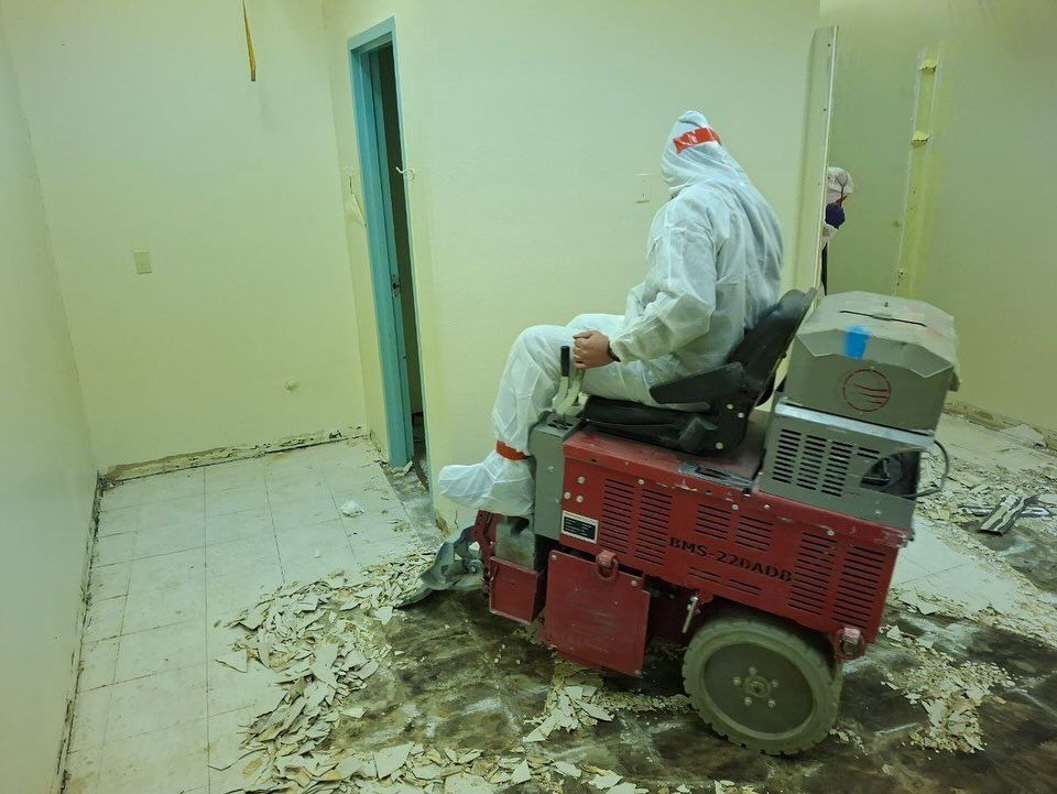 Person in protective suit operating floor removal machine, removing tiles in room.