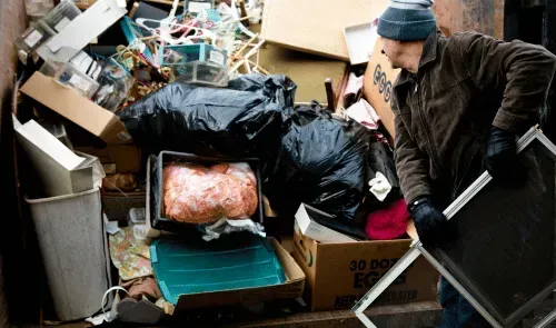 Man loading various household items and black trash bags into a bin.