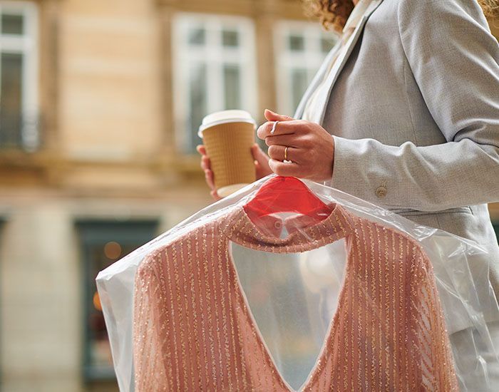 Person holding a coffee and a garment in a dry-cleaning bag outdoors