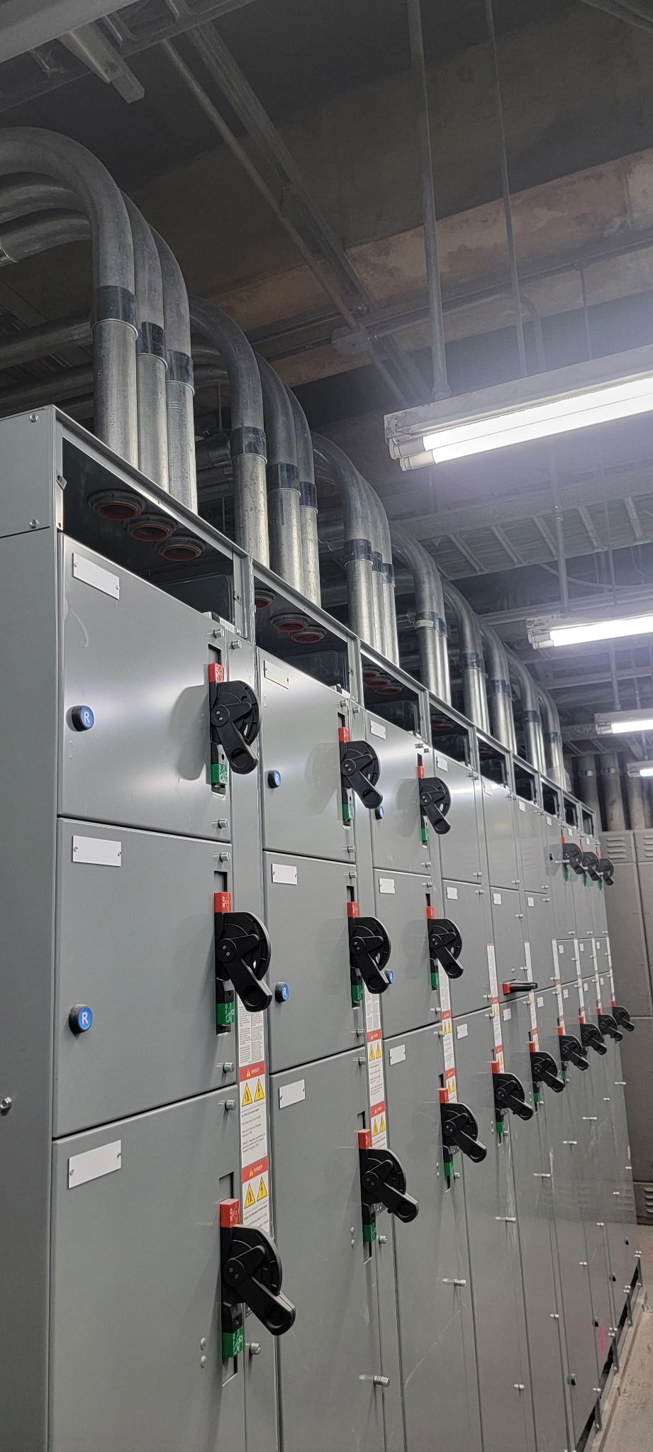Row of gray electrical control cabinets in an industrial facility with red and green indicator lights under fluorescent lighting