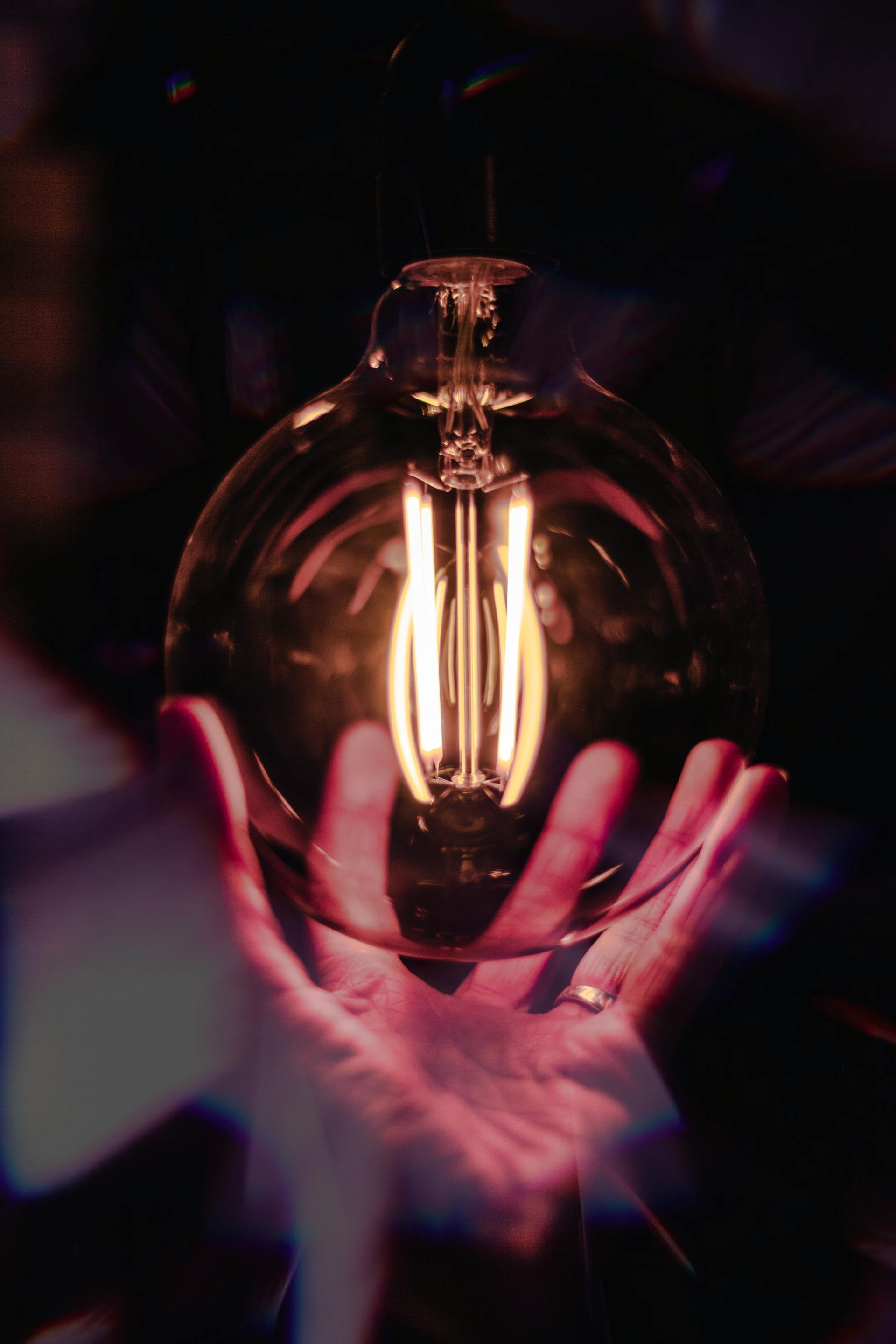 Hand cupping a glowing, vintage-style lightbulb, warm light against a dark background.