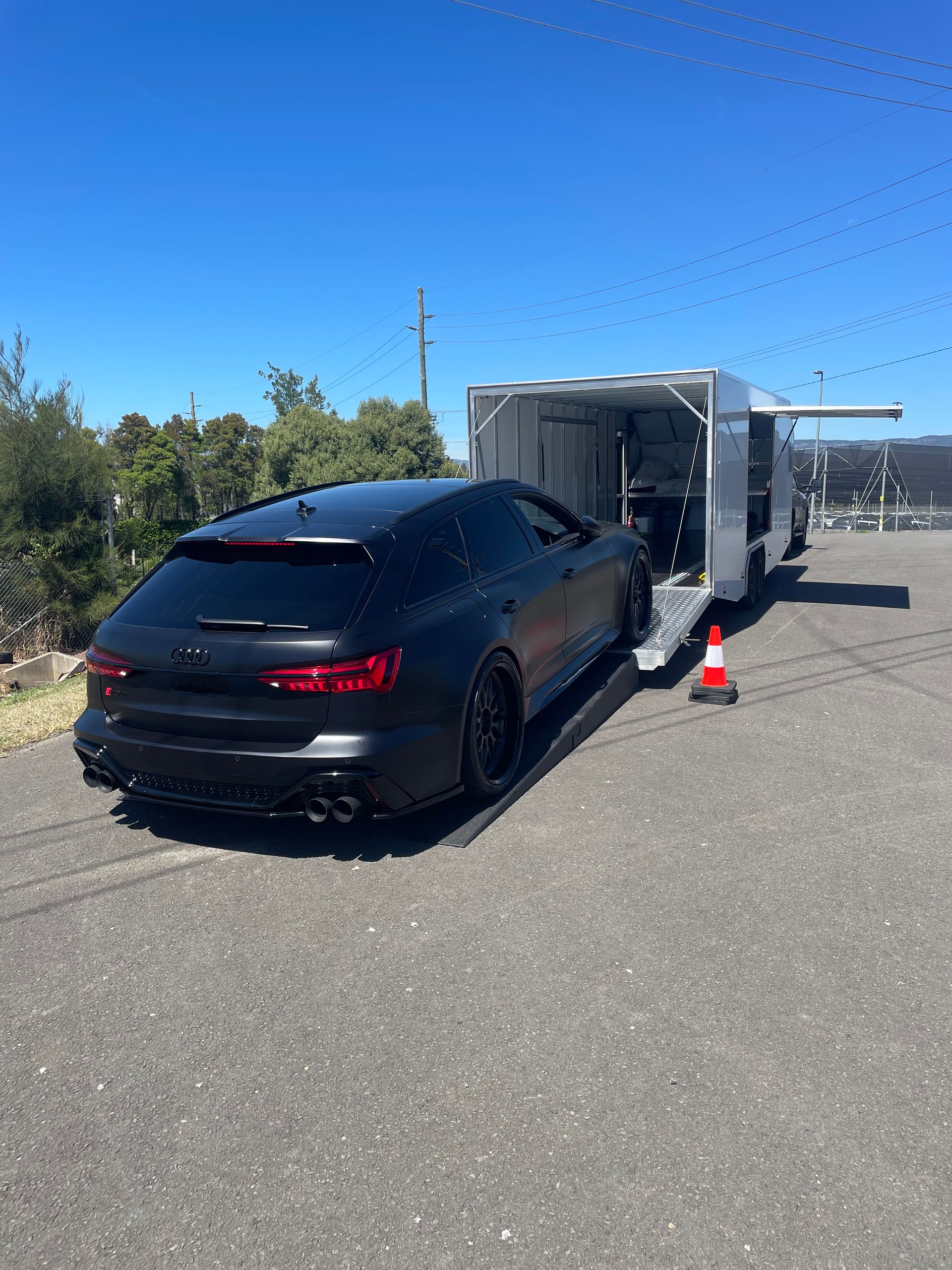Black sedan hitched to a trailer on a sunny roadside, with traffic cones nearby. - Full Throttle Enclosed Car Transport in Kilmore, VIC
