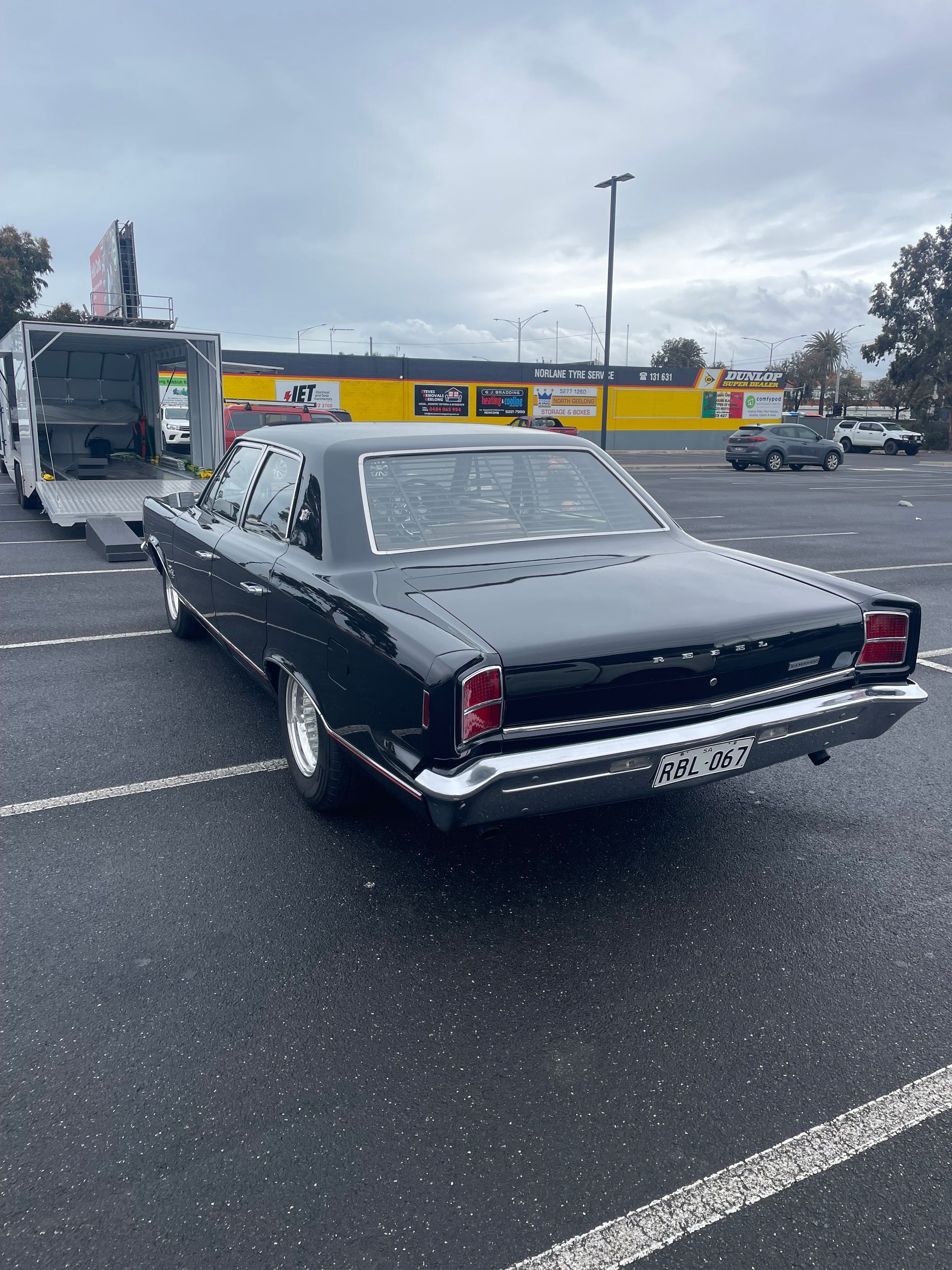 Black classic car parked in a wet lot near a bus, with a cloudy sky overhead - Full Throttle Enclosed Car Transport in Kilmore, VIC