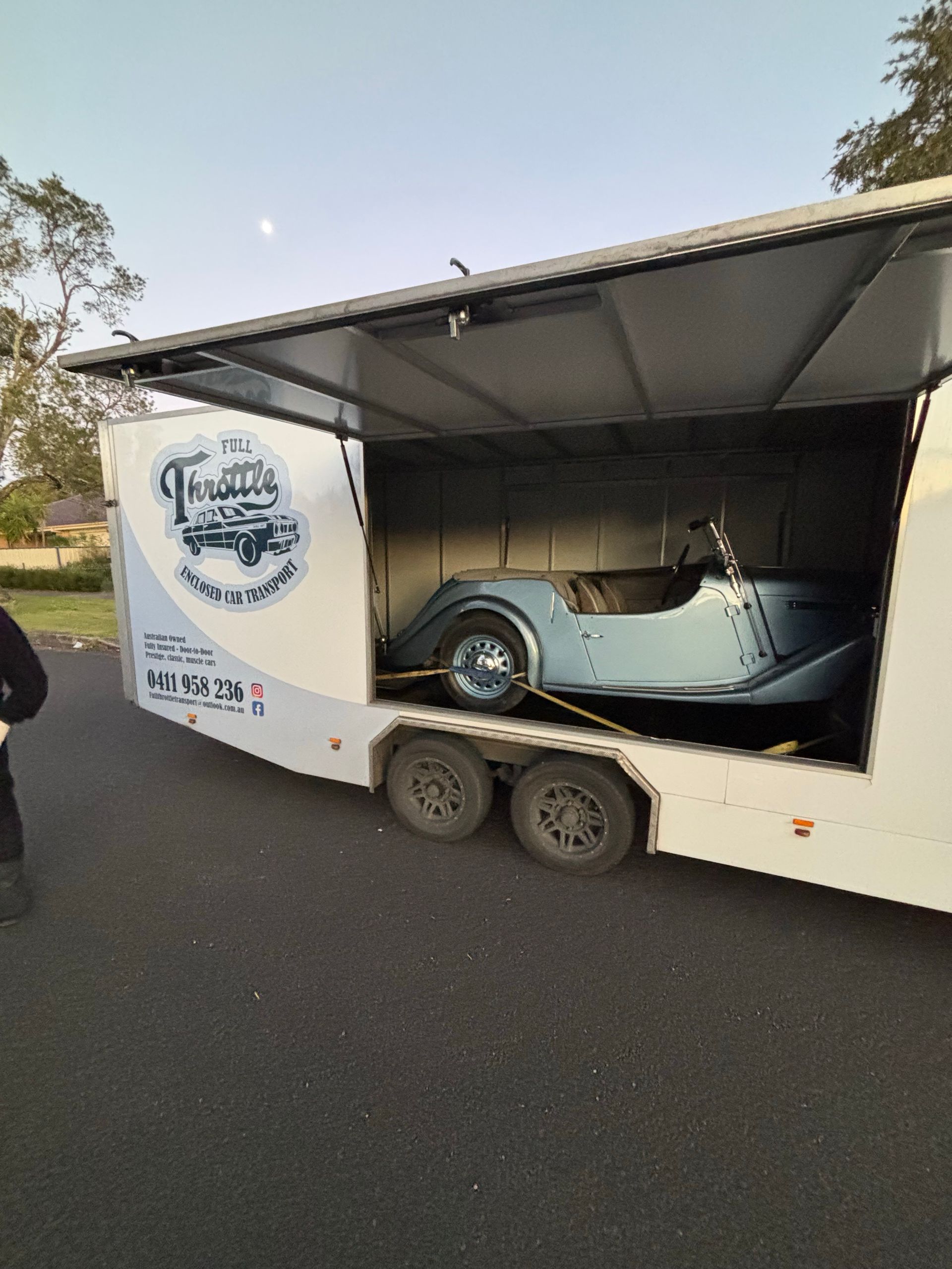 Light blue vintage car inside an open trailer on a road, with a logo on the trailer side. - Full Throttle Enclosed Car Transport in Kilmore, VIC