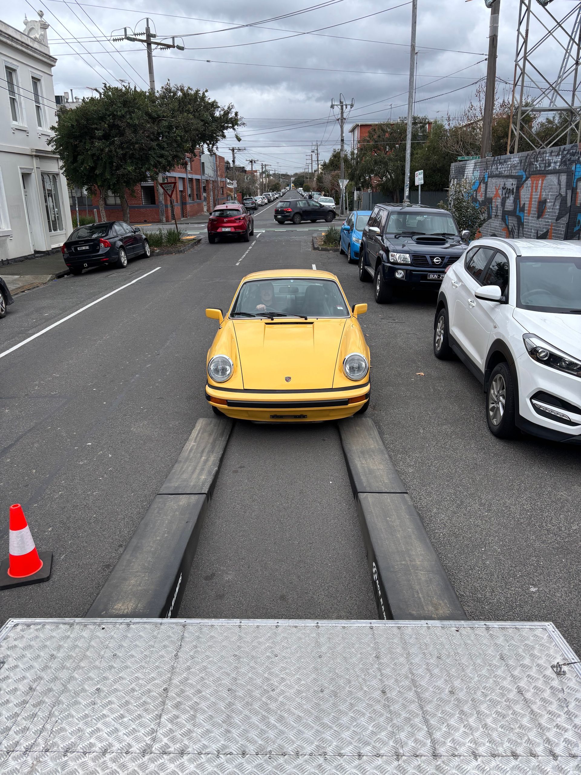 Yellow vintage car on a city street, viewed from the front, with parked cars and overcast sky. - Full Throttle Enclosed Car Transport in Kilmore, VIC