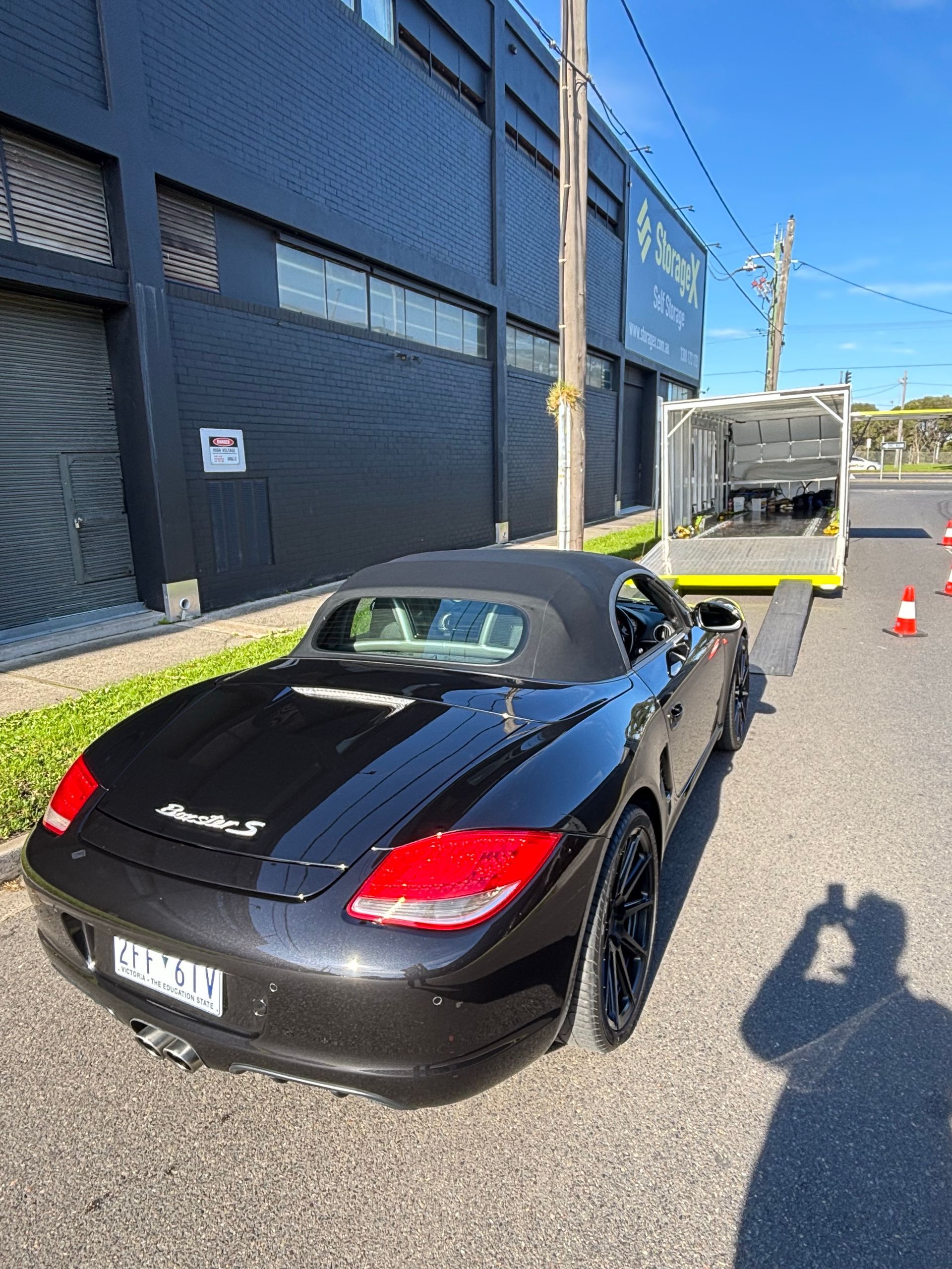 Black sports car parked by a blue building, with cones and a truck in the background. - Full Throttle Enclosed Car Transport in Kilmore, VIC