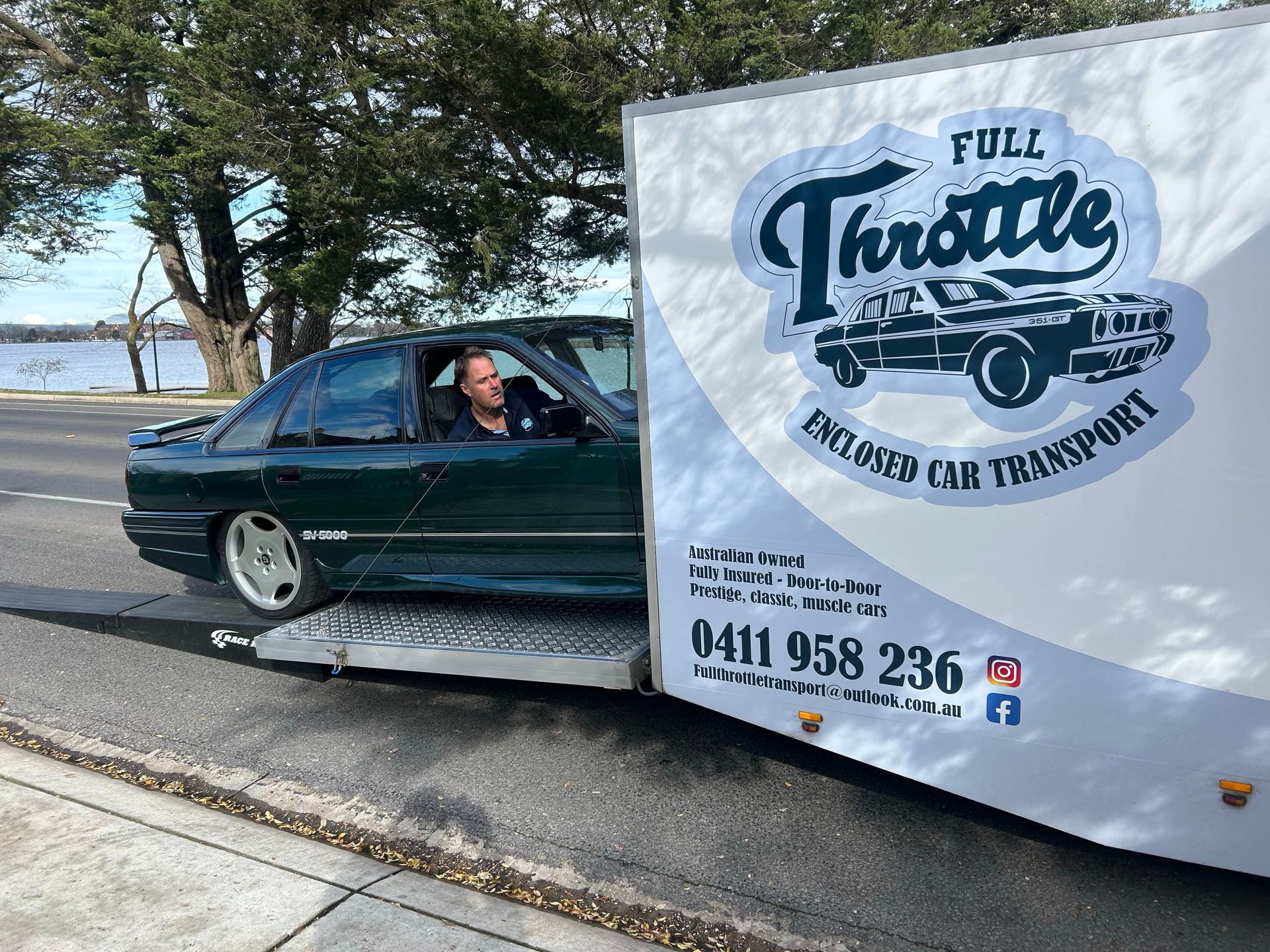 Tow truck hauling a dark green car with a “Full Throttle” sign along a roadside by the water - Full Throttle Enclosed Car Transport in Kilmore, VIC