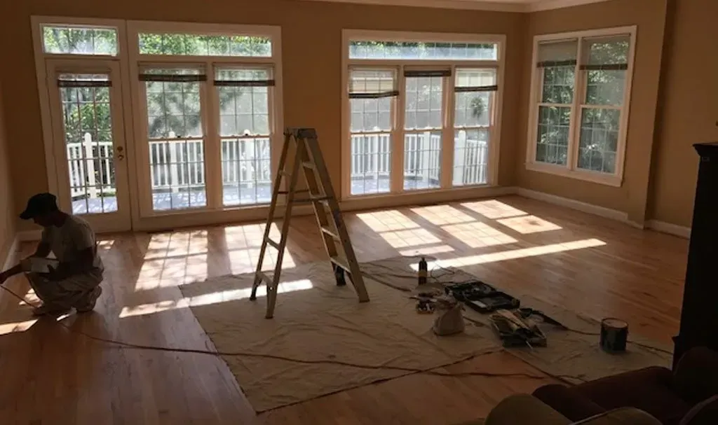 A man is kneeling down in an empty living room with a ladder.
