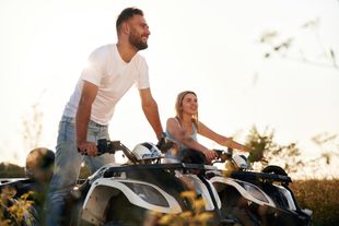 Couple on ATVs, driving outdoors. Man smiles, woman points. Sunny day, field in background.