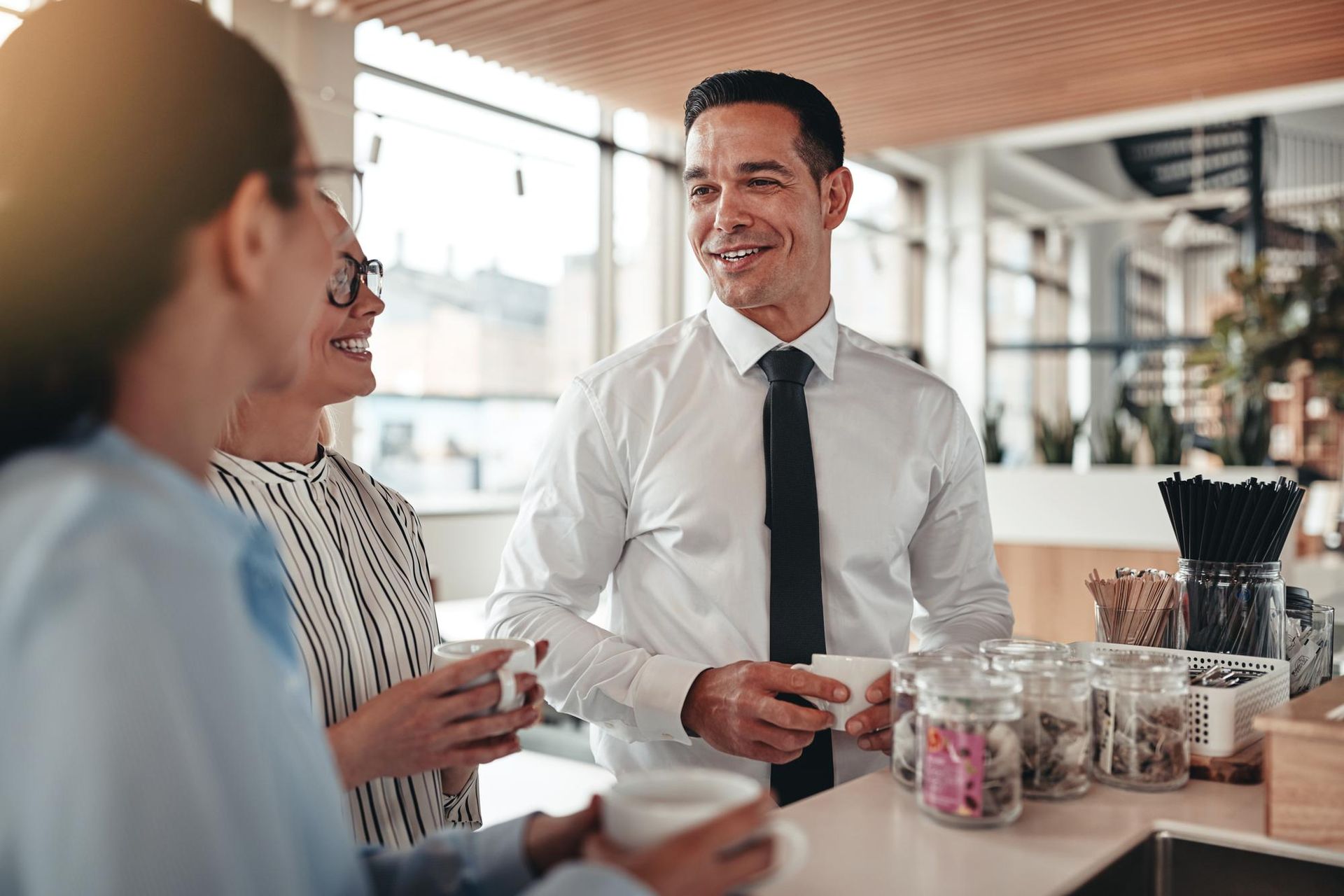 Three people in a brightly lit cafe. A man in a white shirt smiles as he talks to two women holding mugs.