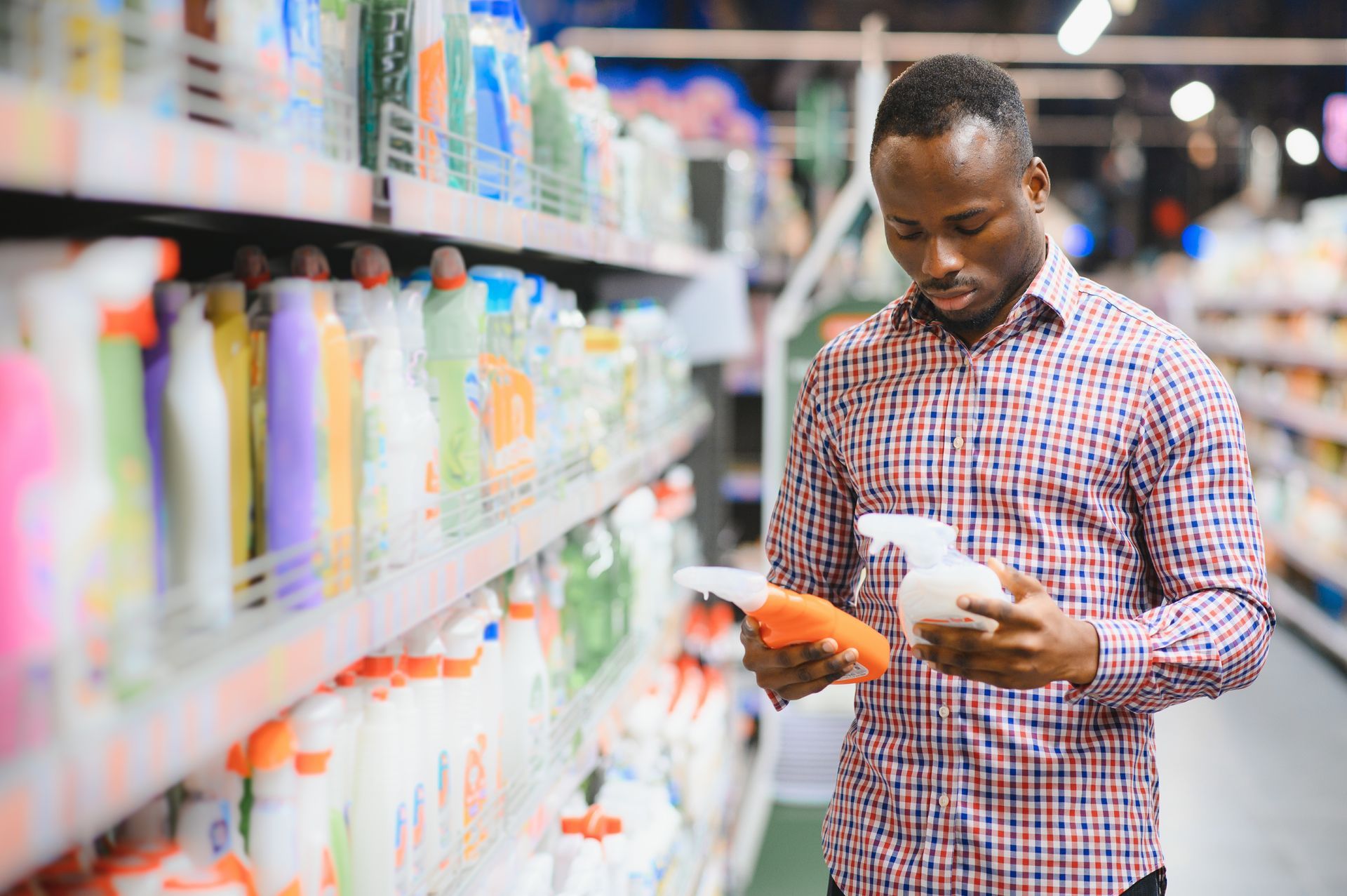 Man in plaid shirt browsing cleaning products in a store, comparing bottles.