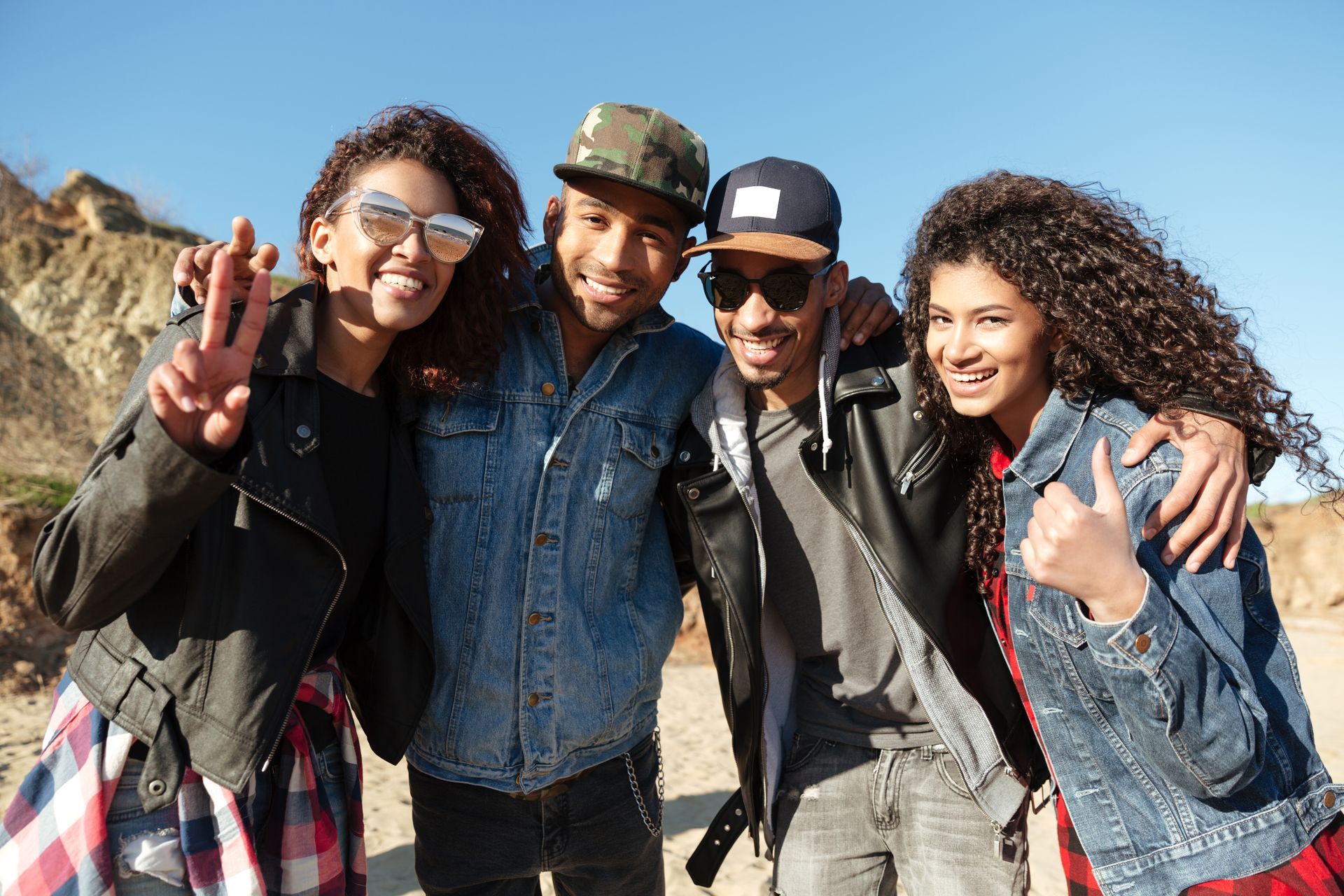 Four friends with arms around each other, smiling, outside. Two wear denim jackets, one gives peace sign.