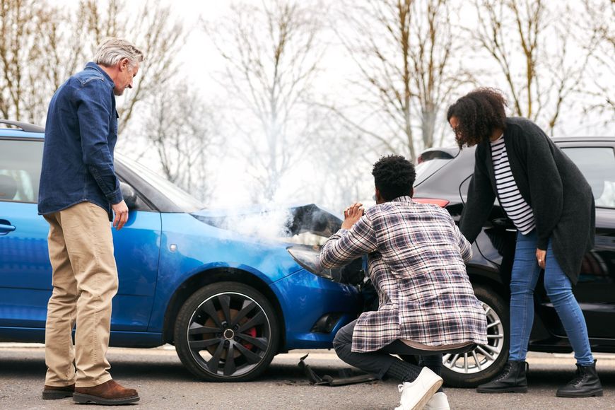 People examine a damaged blue car with smoke after an accident.