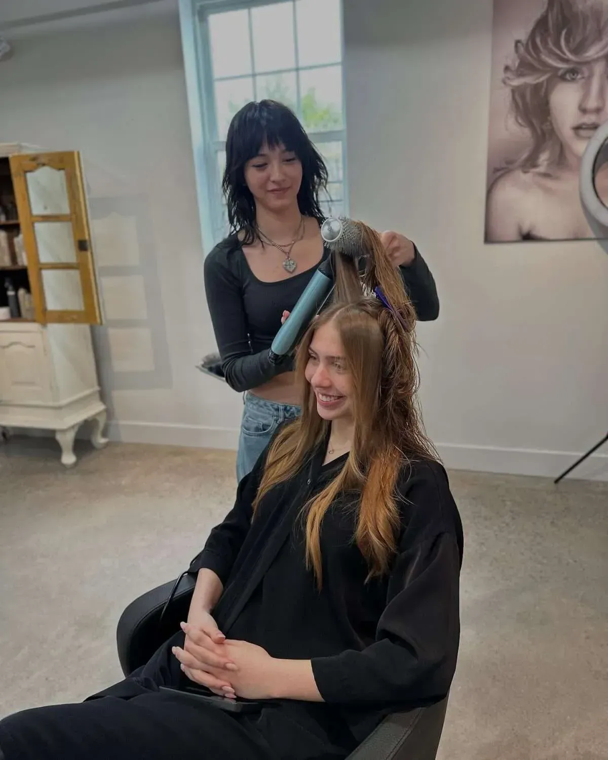 Hair stylist curling a seated client’s hair in a salon; both wearing dark tops.