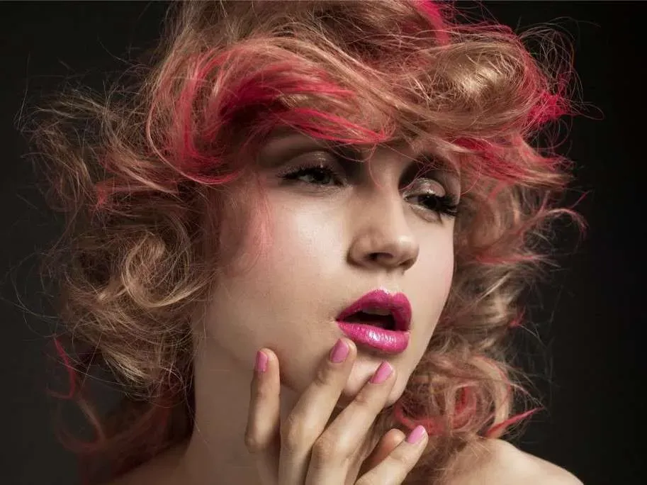 Close-up portrait of a person with curly hair, pink makeup, and hand near face against a dark background