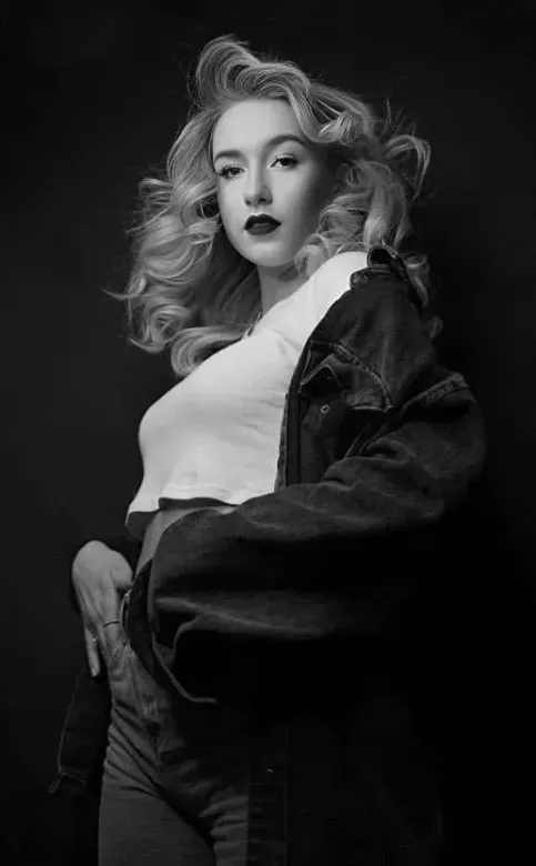 Black-and-white portrait of a person with wavy hair wearing a light shirt and dark jacket, posing confidently.