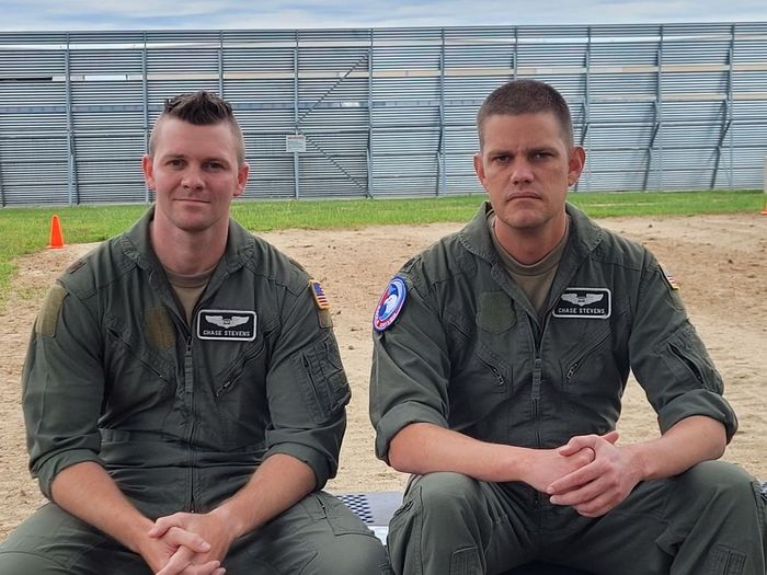 Two men in green flight suits sit outside; one has a mohawk and smiles.