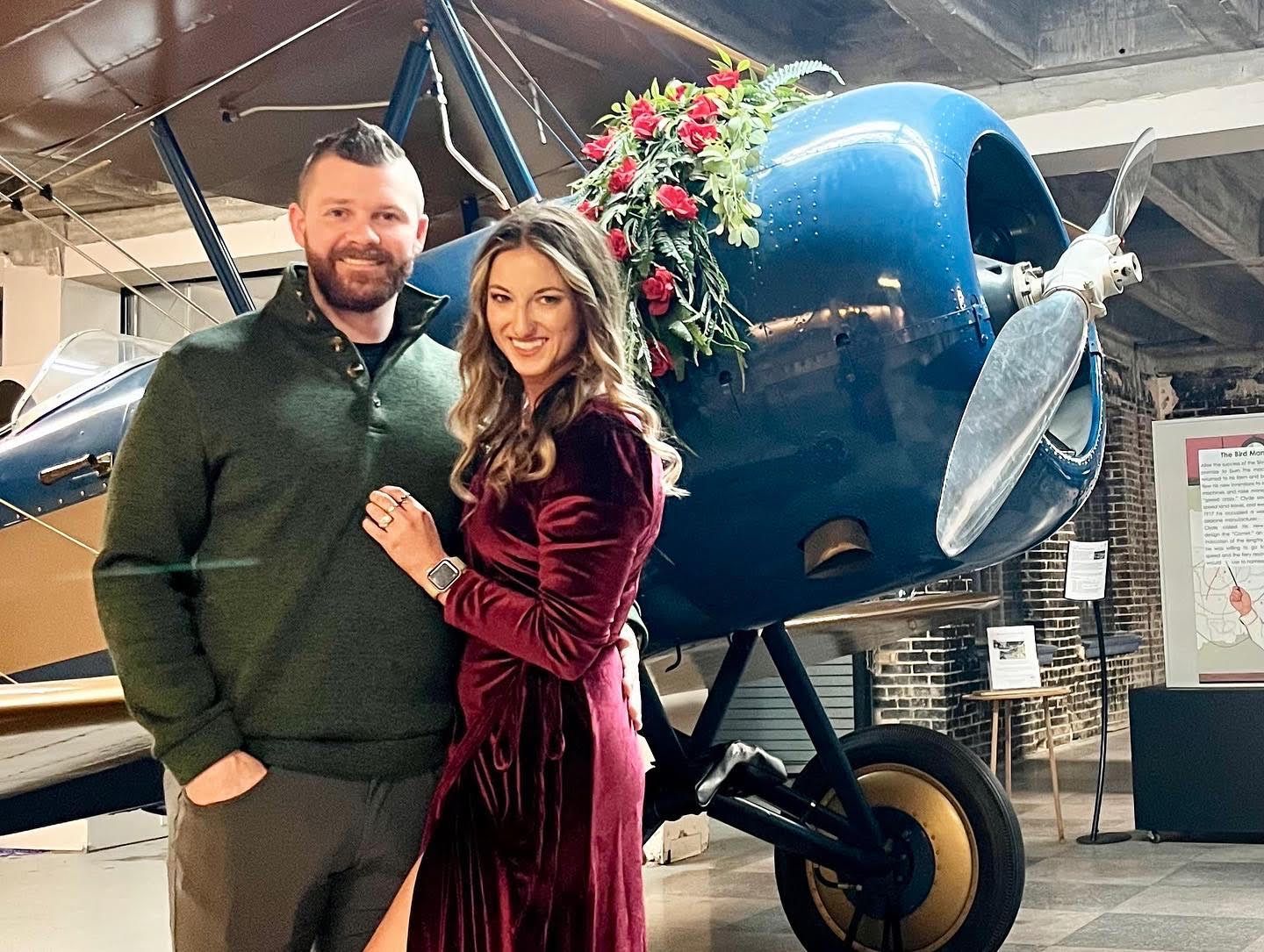 Couple posing in front of a blue biplane adorned with flowers, in a museum.