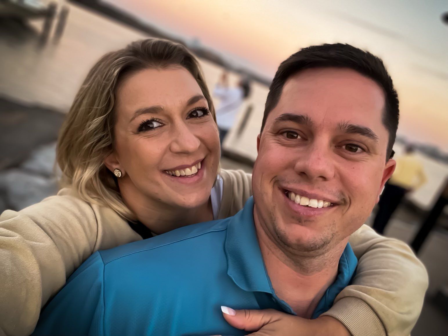 Couple smiling at camera, woman hugging man from behind. Sunset over water in background.