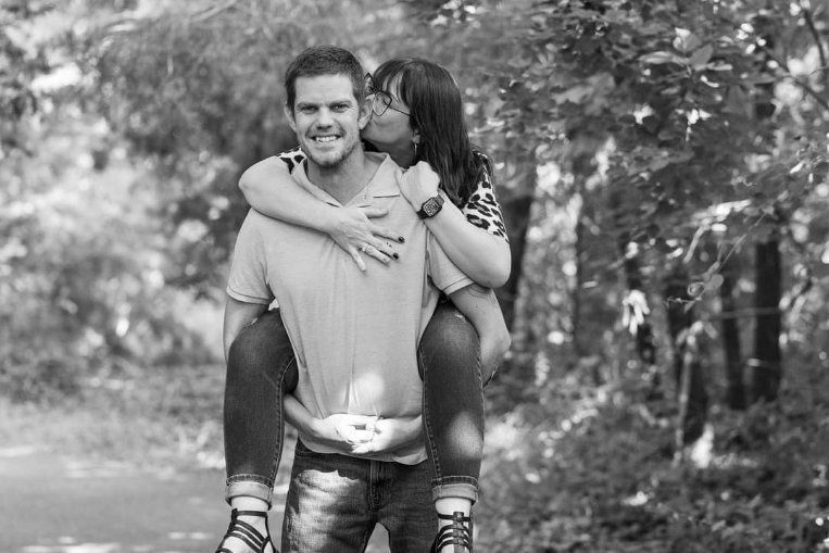 Man giving woman a piggyback ride, kissing her cheek. Outdoors with trees in black and white.