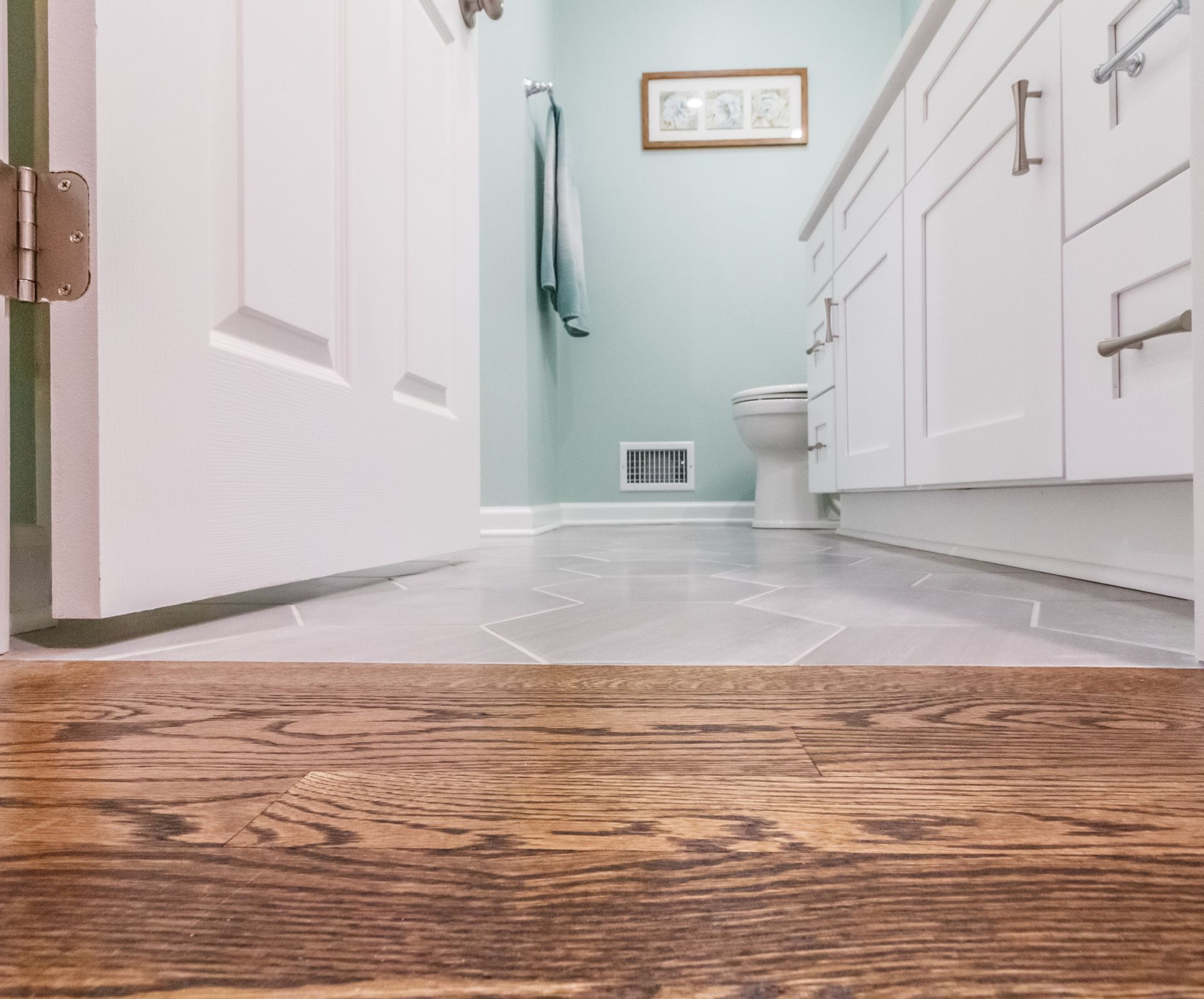 Low-angle view of a bright bathroom with white cabinets, pale walls, and wood flooring in the foreground