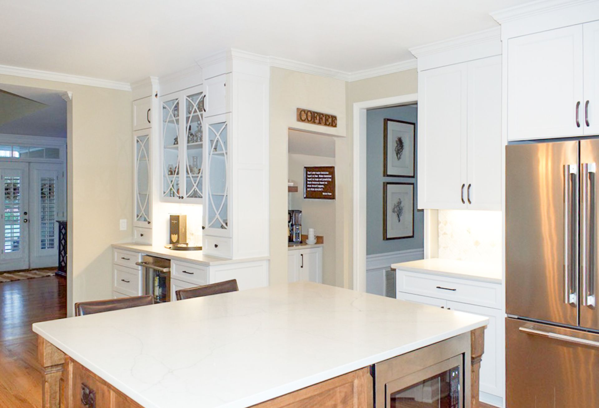 Bright white kitchen with a large island, stainless steel fridge, and built-in cabinets.