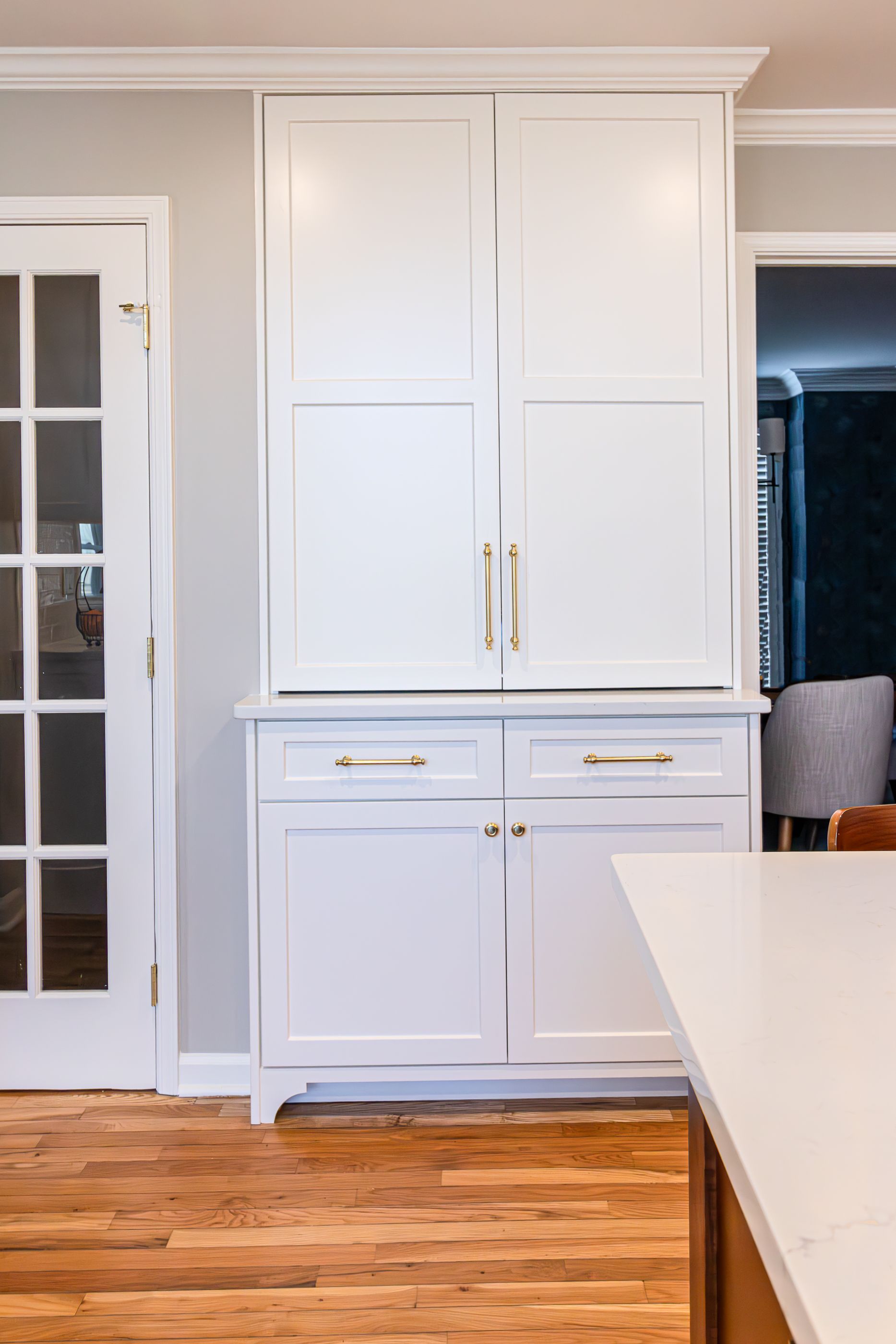 White built-in kitchen cabinets with gold handles beside a glass door and white countertop in a bright room