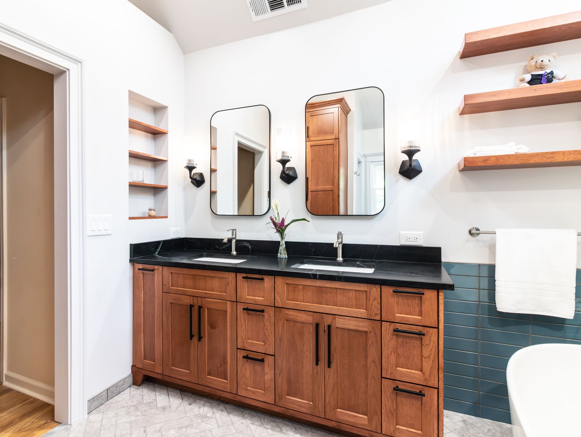 Bathroom vanity with wood cabinets, black countertop, mirrors, wall sconces, and teal tile backsplash