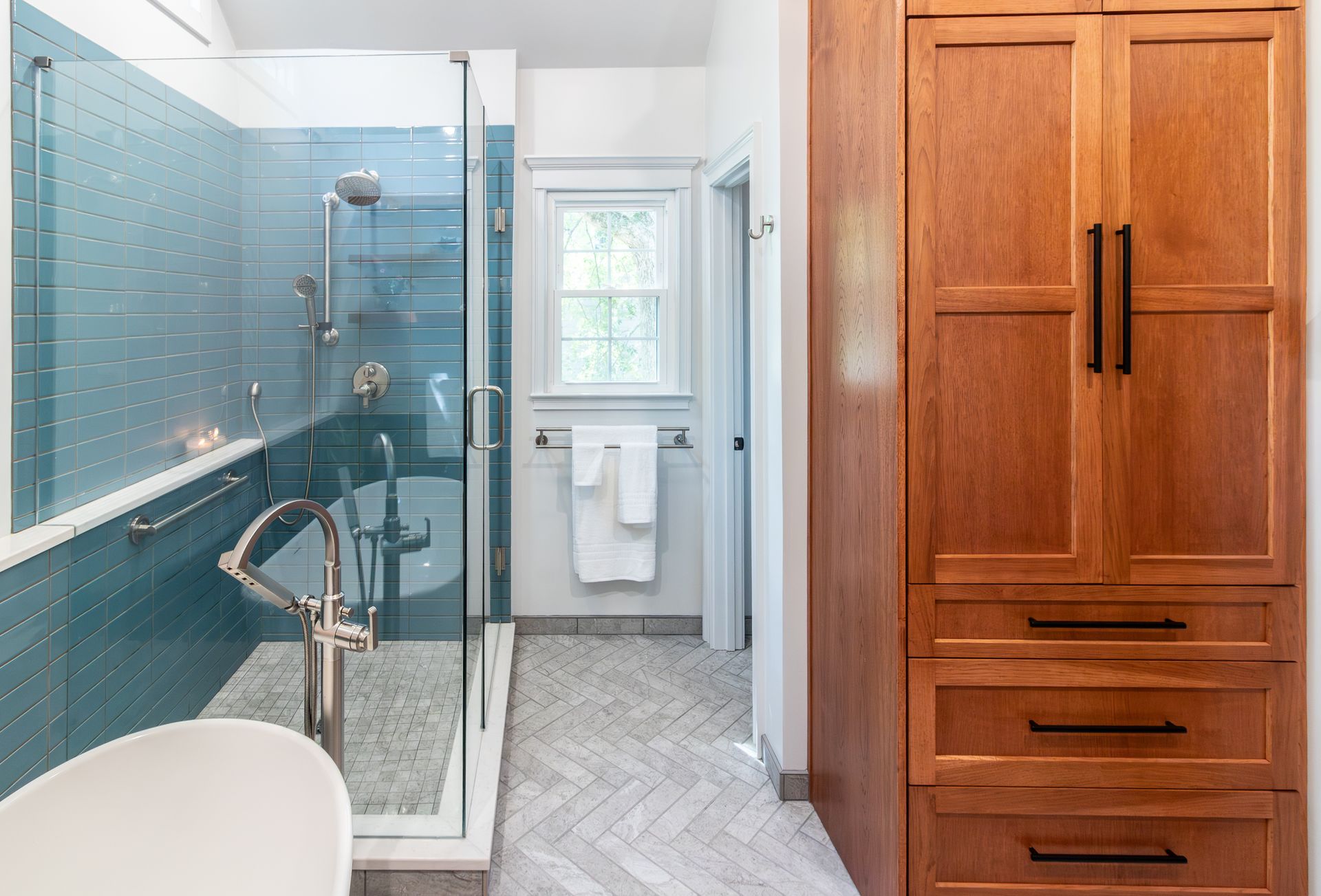 Bright bathroom with glass shower, blue tiled wall, small sink, window, and wooden cabinet