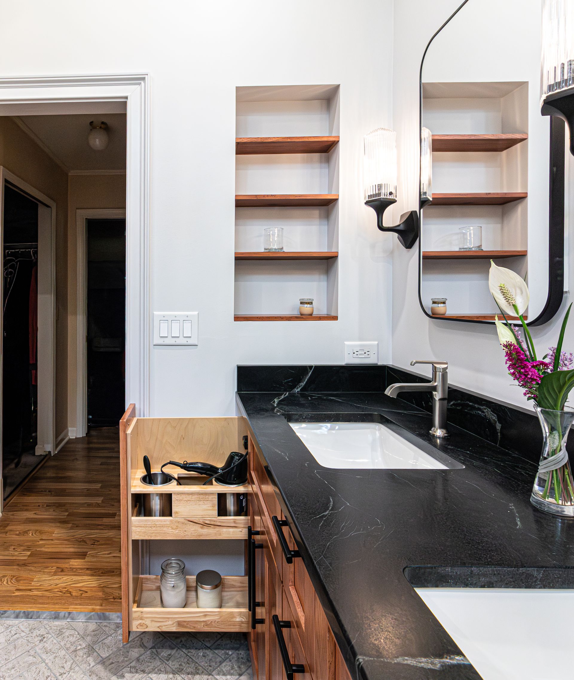 Bathroom vanity with black countertop, white sink, wall shelves, and wooden storage cart near doorway