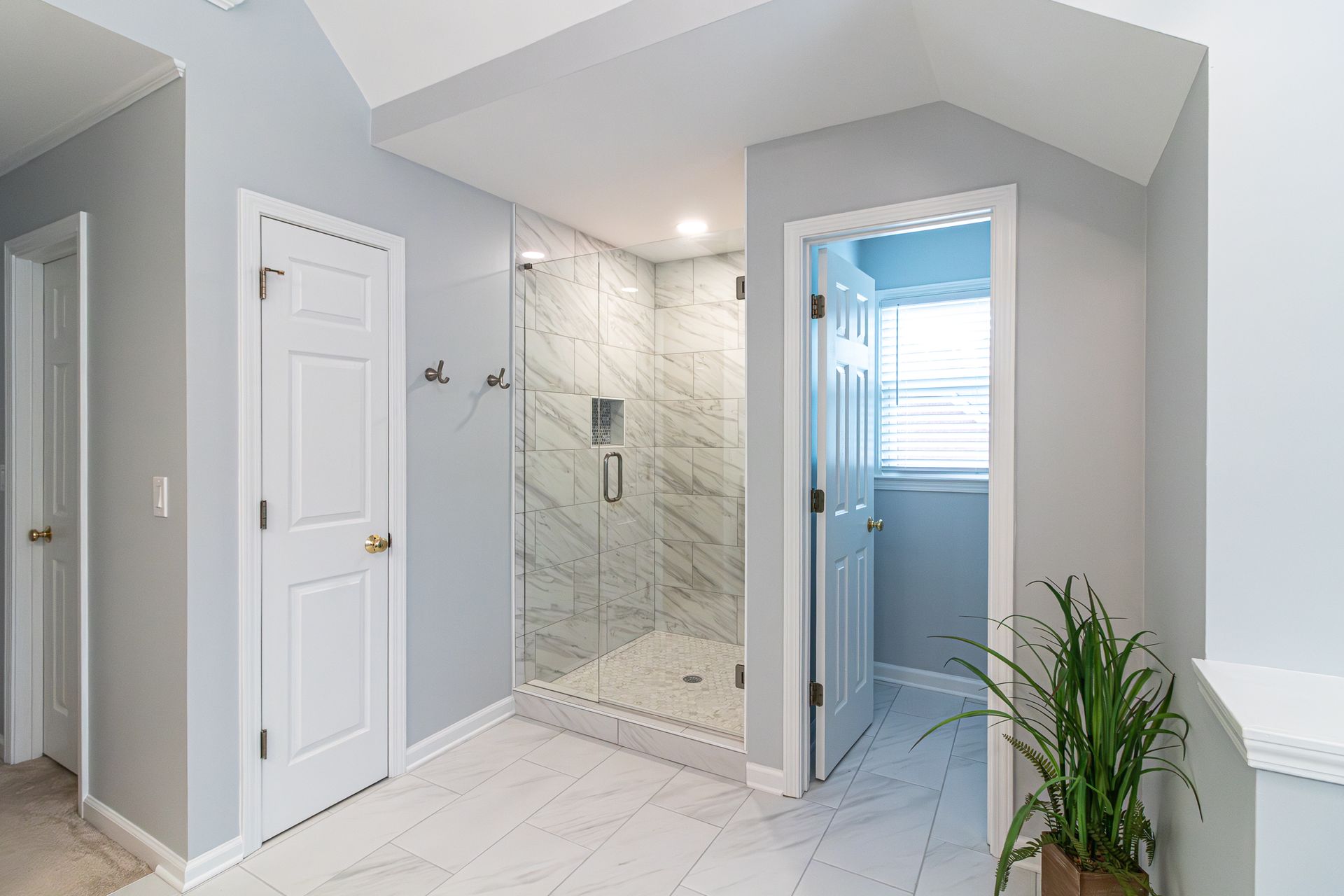 Bright bathroom with a glass shower, white tile, and a potted plant by the doorway.